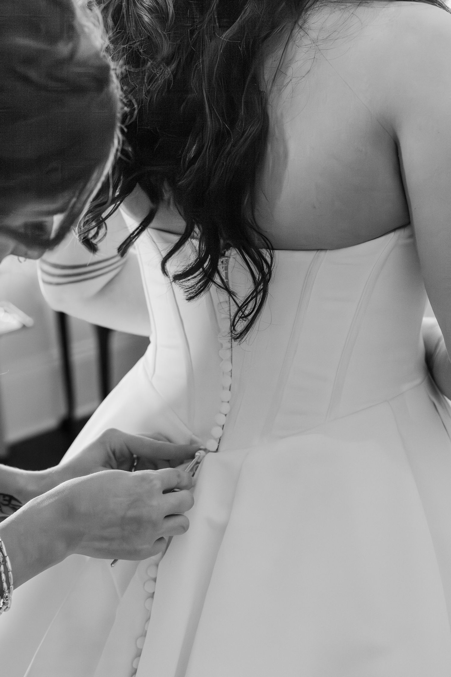 Black and white photo of a bride getting her wedding dress buttoned up the back by a bridesmaid.