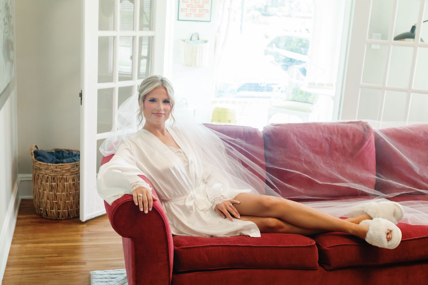 Bride in white robe and veil relaxing on a red velvet sofa, wearing fluffy slippers in a bright, elegant room.