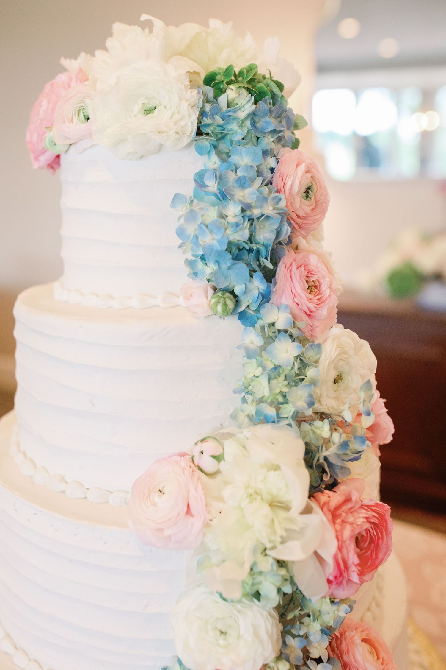 Four-tier white wedding cake adorned with cascading pink ranunculus, blue hydrangeas, and white flowers.