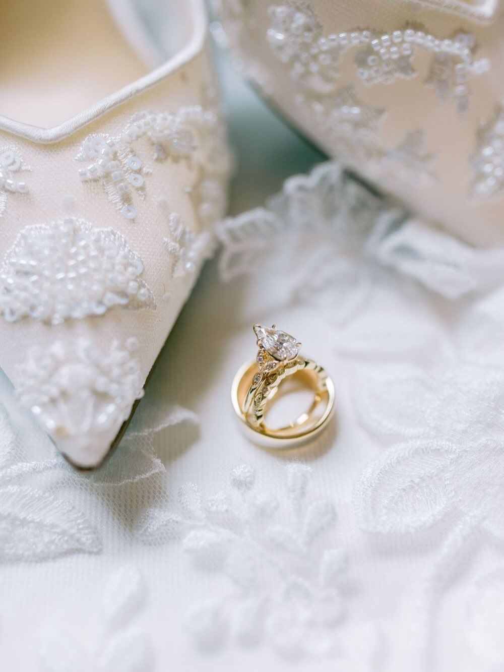 Gold wedding bands and diamond engagement ring rest against white lace bridal shoes and fabric.