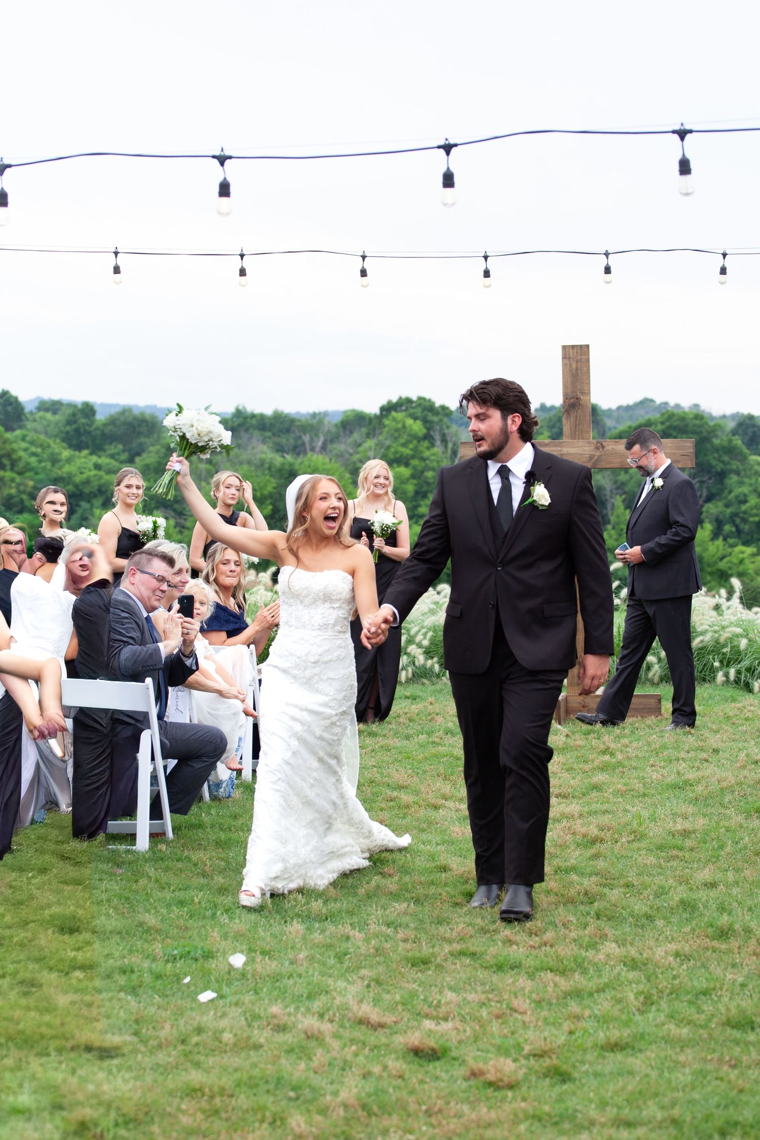 Joyful bride and groom walk down outdoor aisle after wedding ceremony, guests cheering, string lights overhead.