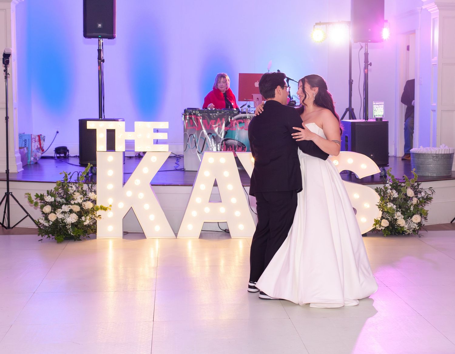 Bride and groom share first dance in front of illuminated marquee letters at elegant wedding reception.
