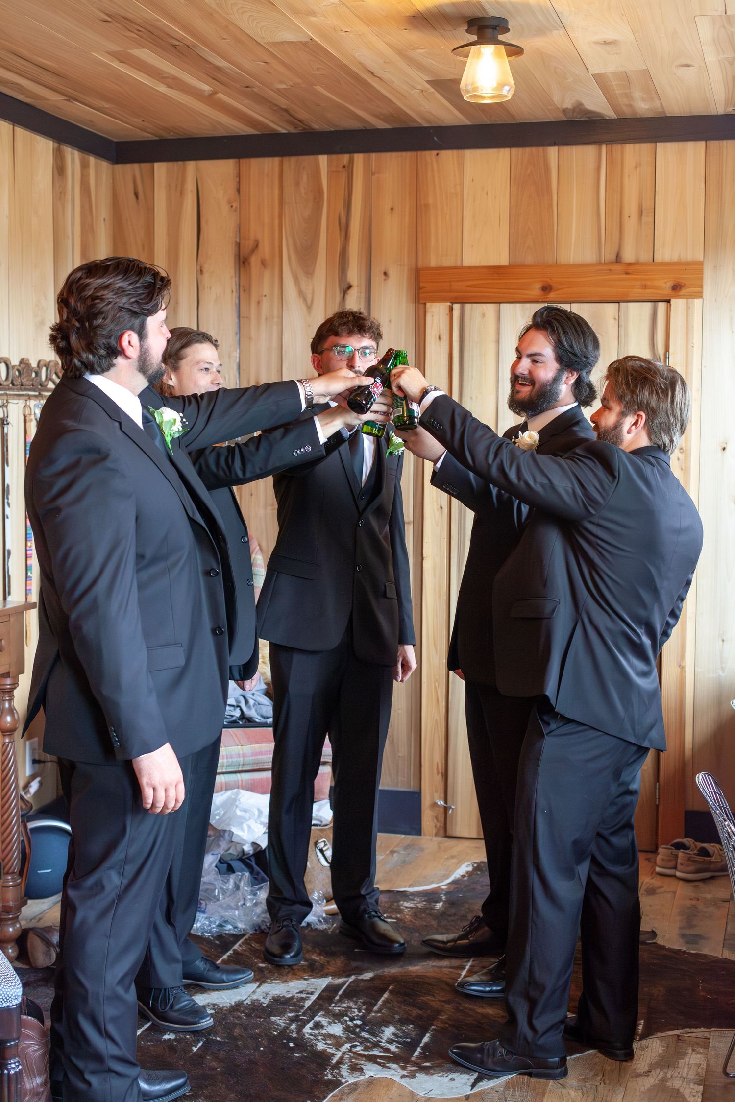 Groomsmen in black suits toasting with beer bottles in a rustic wood-paneled room before a wedding ceremony.