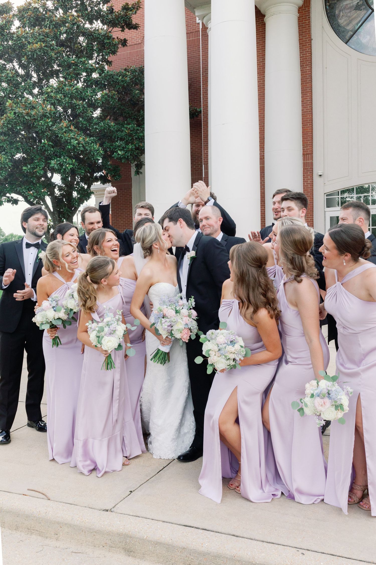 Wedding party poses outside brick church, bride and groom kissing surrounded by bridesmaids in lavender gowns and groomsmen in tuxedos.