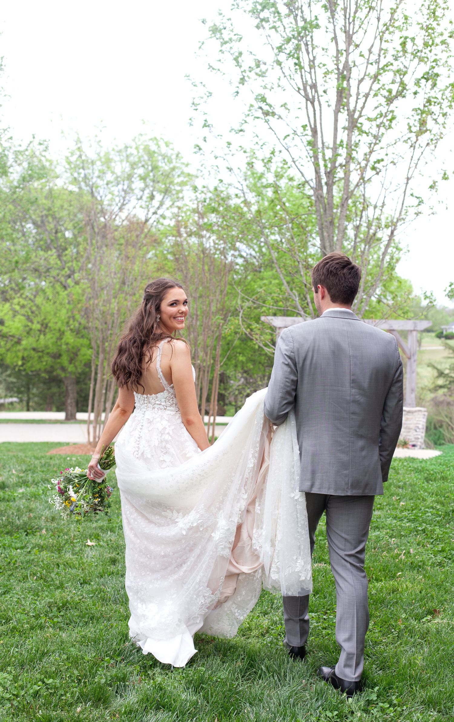 Bride in lace gown holds bouquet as couple walks hand-in-hand across green lawn on wedding day.