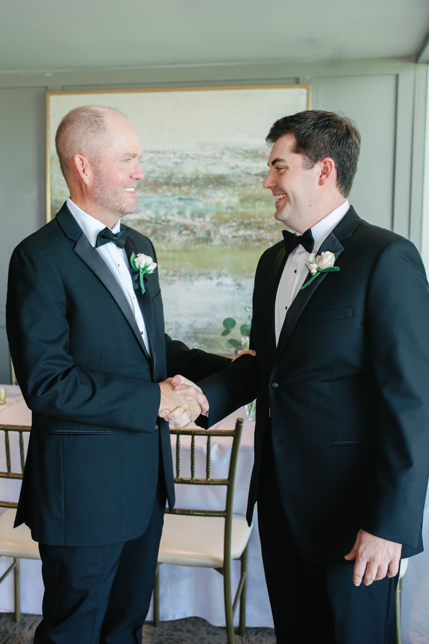 Two men in black tuxedos with boutonnieres shaking hands and smiling at a formal wedding event.