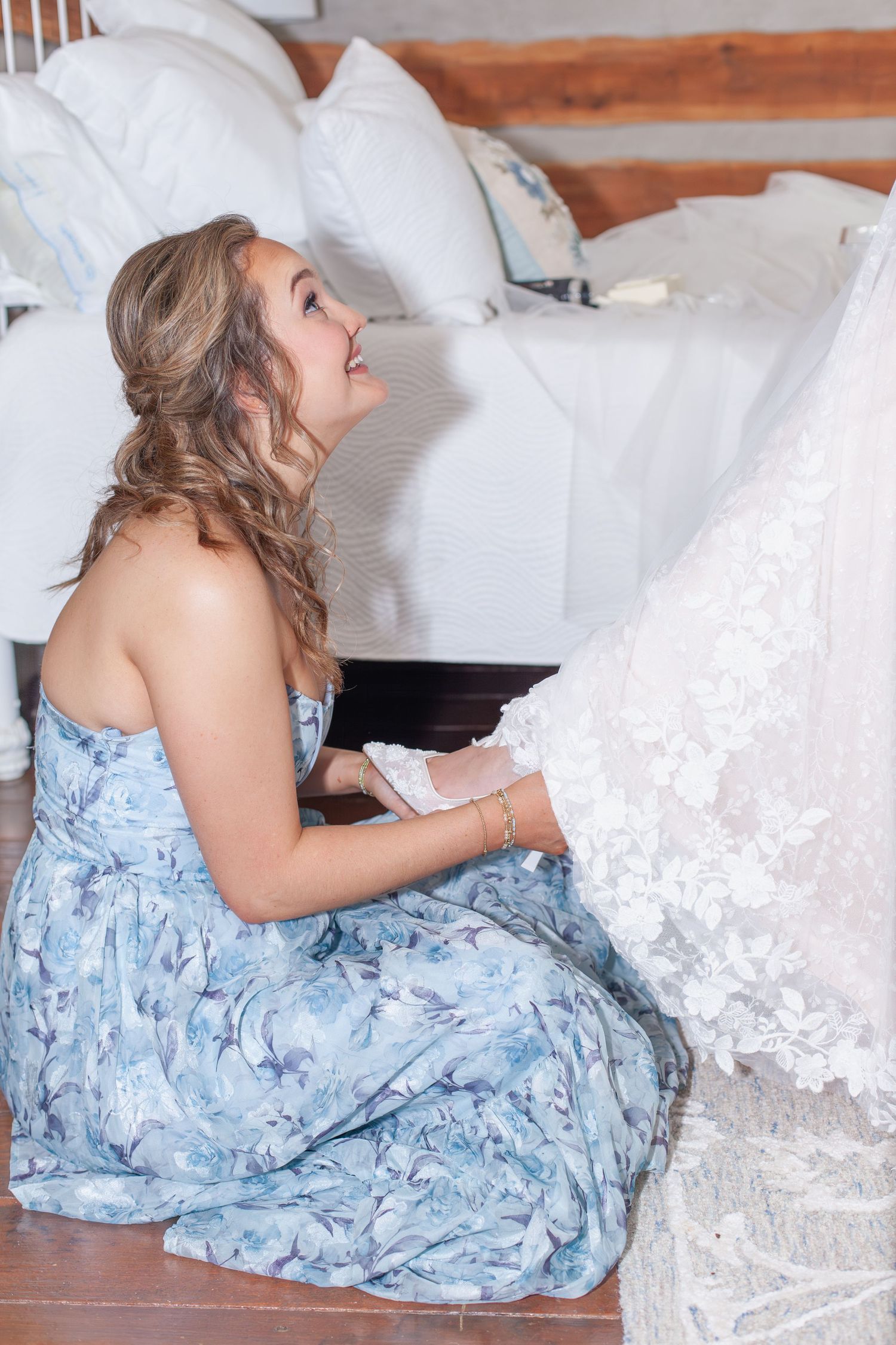 Bridesmaid in blue floral dress kneeling beside a white lace wedding gown indoors.