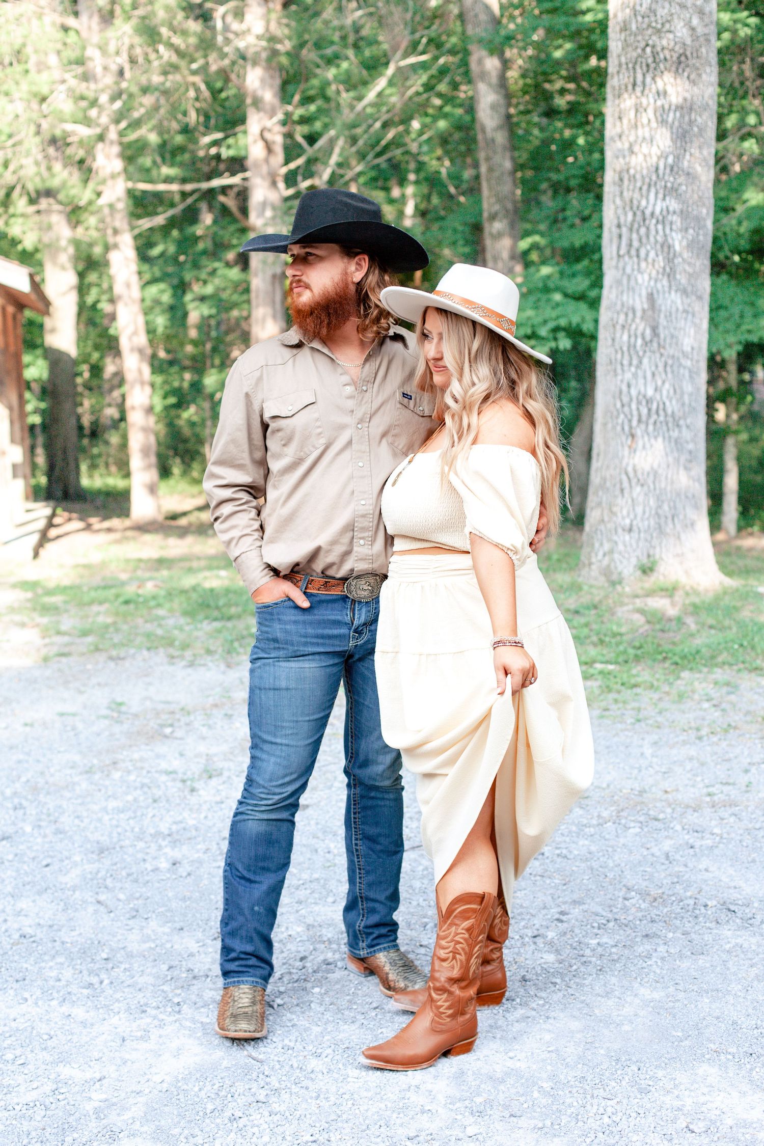 A romantic couple in western attire shares tender moments on a gravel path surrounded by tall trees in a forest setting.
