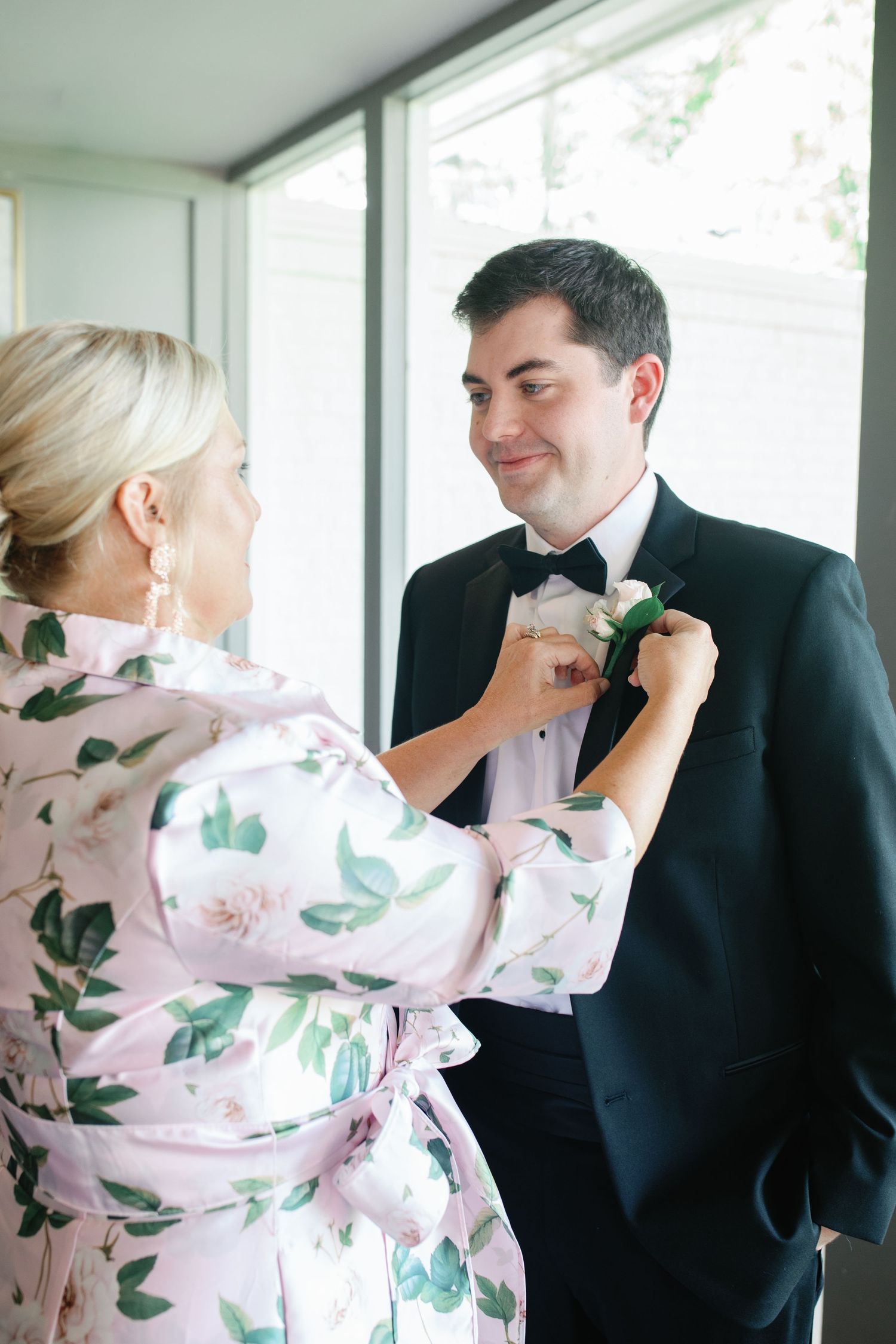 Woman in floral robe pinning boutonniere on groom in black tuxedo before wedding ceremony.