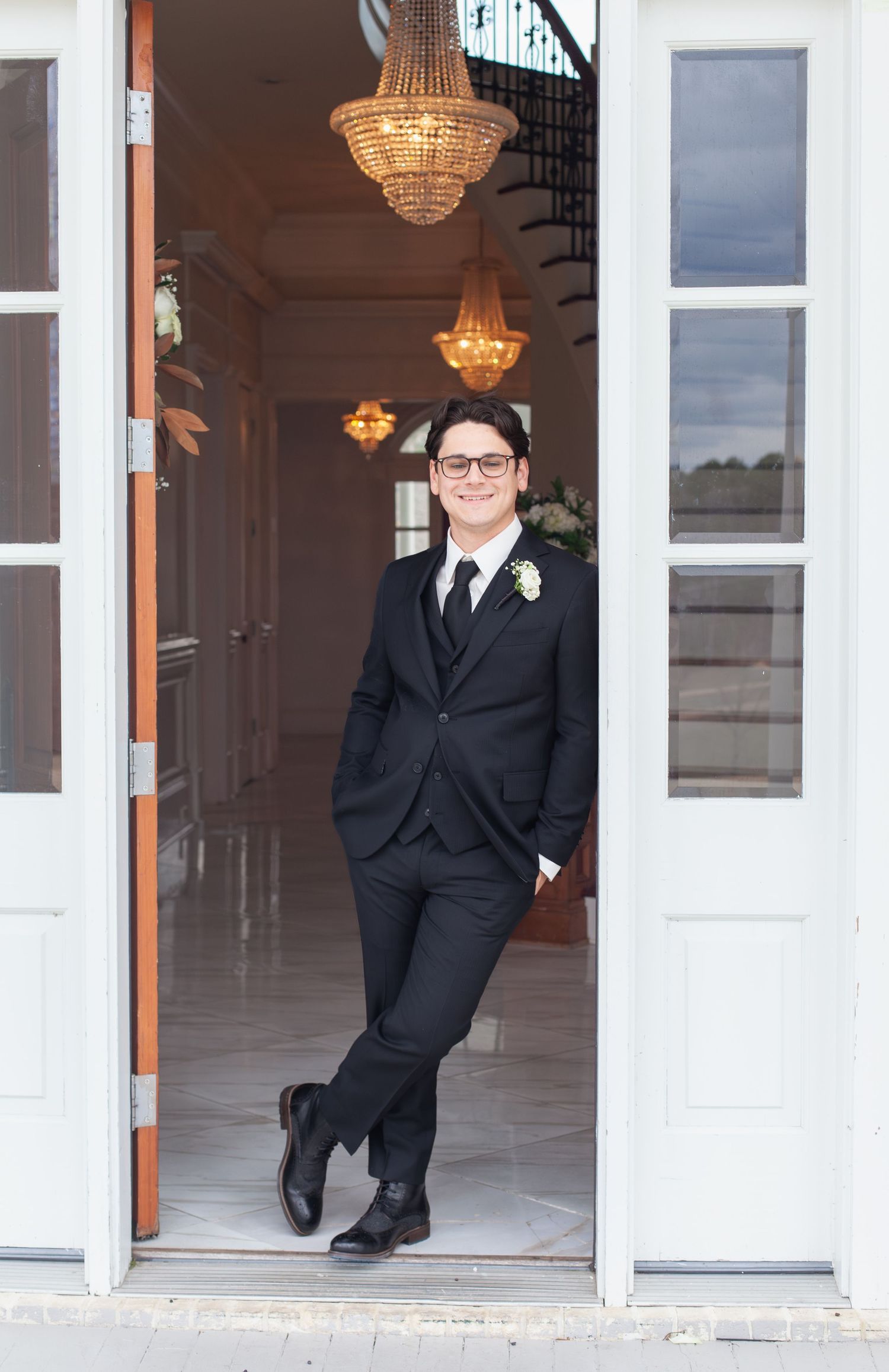 Dapper groom in black suit with boutonniere leaning against open French doors beneath elegant crystal chandeliers.