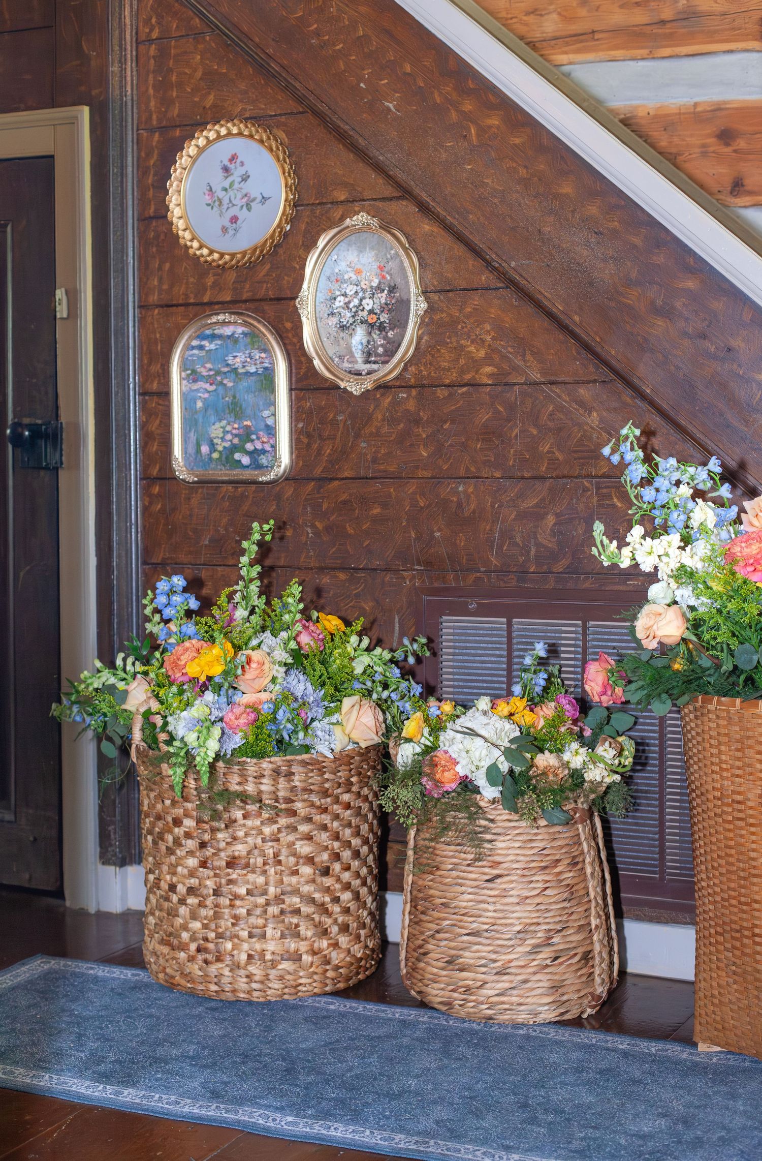 Wicker baskets filled with colorful spring flowers arranged near a wooden staircase with framed floral artwork on the wall.