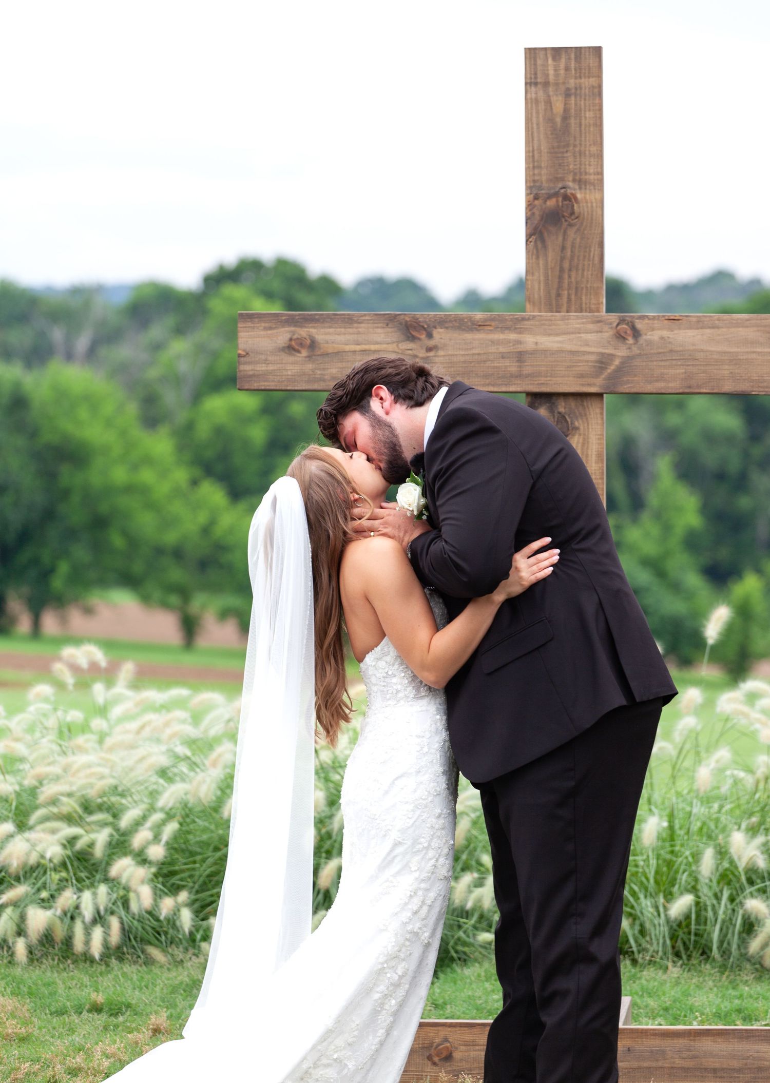 Bride and groom share first kiss at outdoor wedding ceremony in front of rustic wooden cross surrounded by lush greenery.