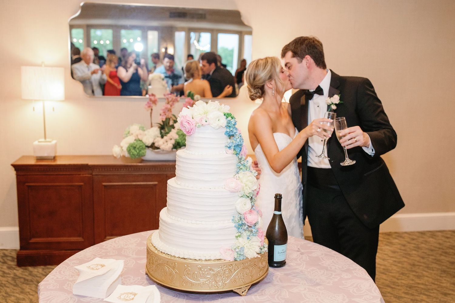 Bride and groom kissing behind a white four-tier wedding cake with floral decor and champagne at their reception.