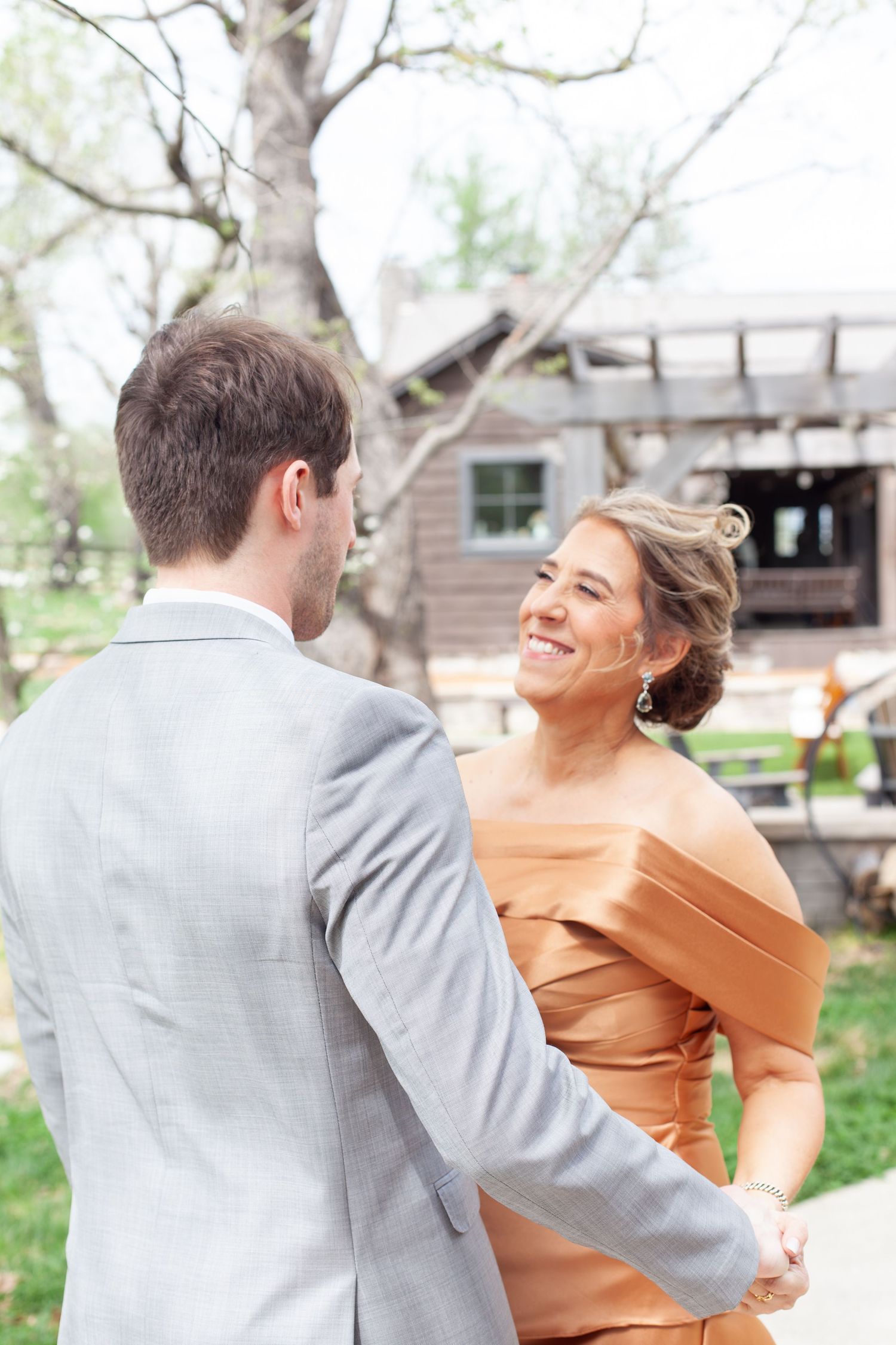 Smiling bride in copper off-shoulder gown holds hands with groom in gray suit outdoors at rustic venue.