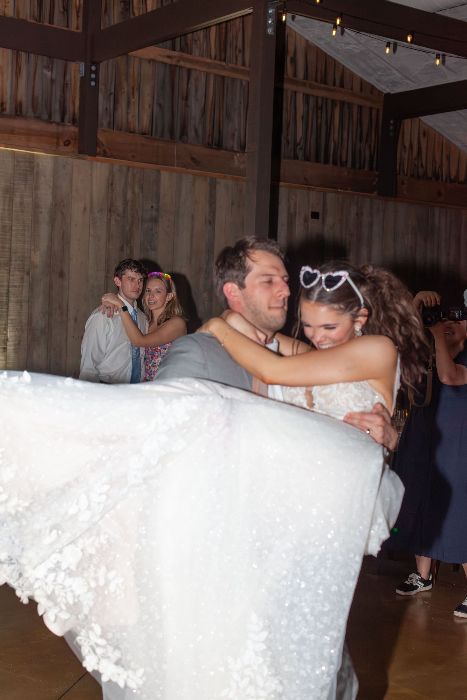 Groom carrying bride in white floral gown on dance floor at rustic barn wedding reception with guests dancing behind them.
