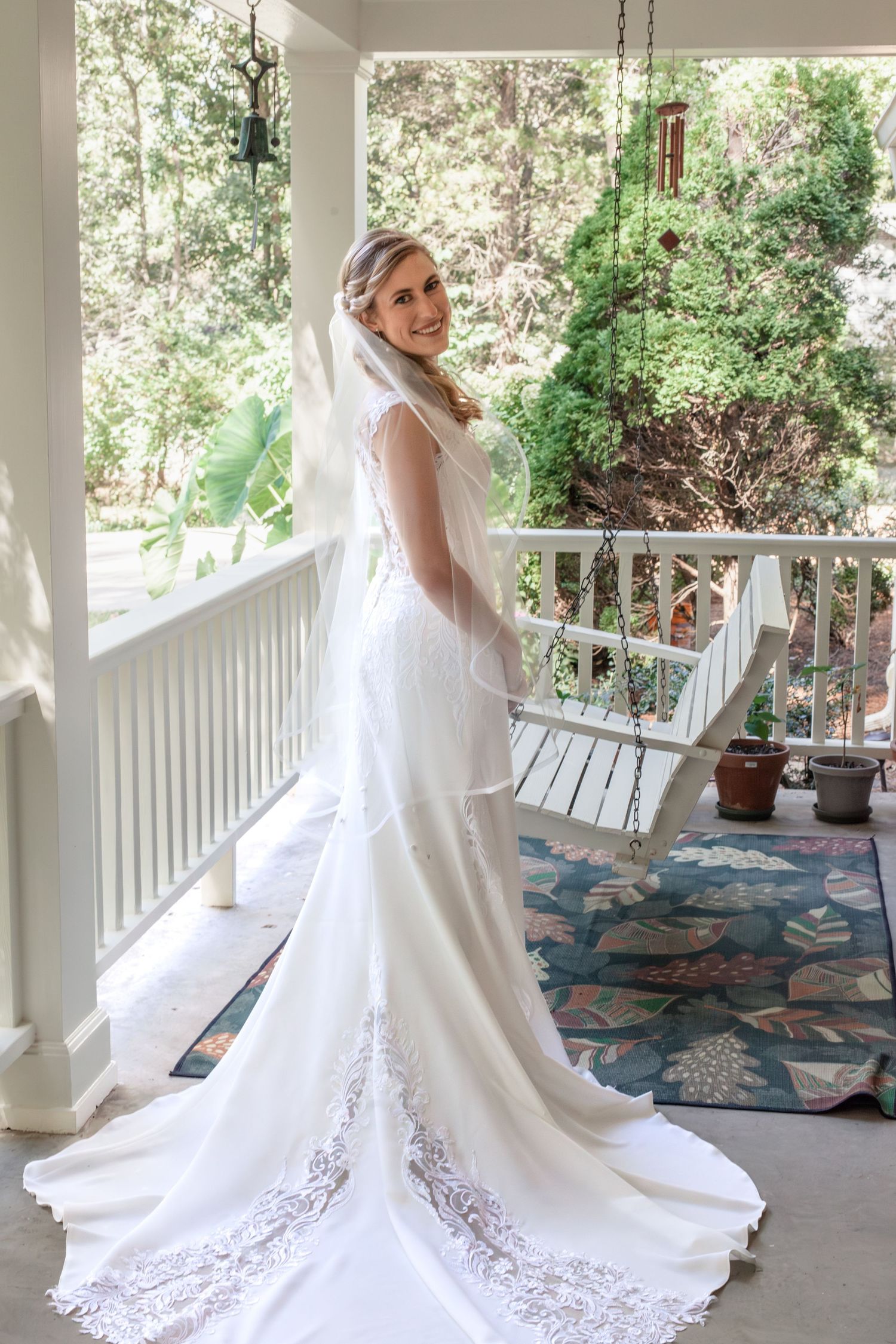 A bride in a lace-detailed wedding gown poses on a covered porch.