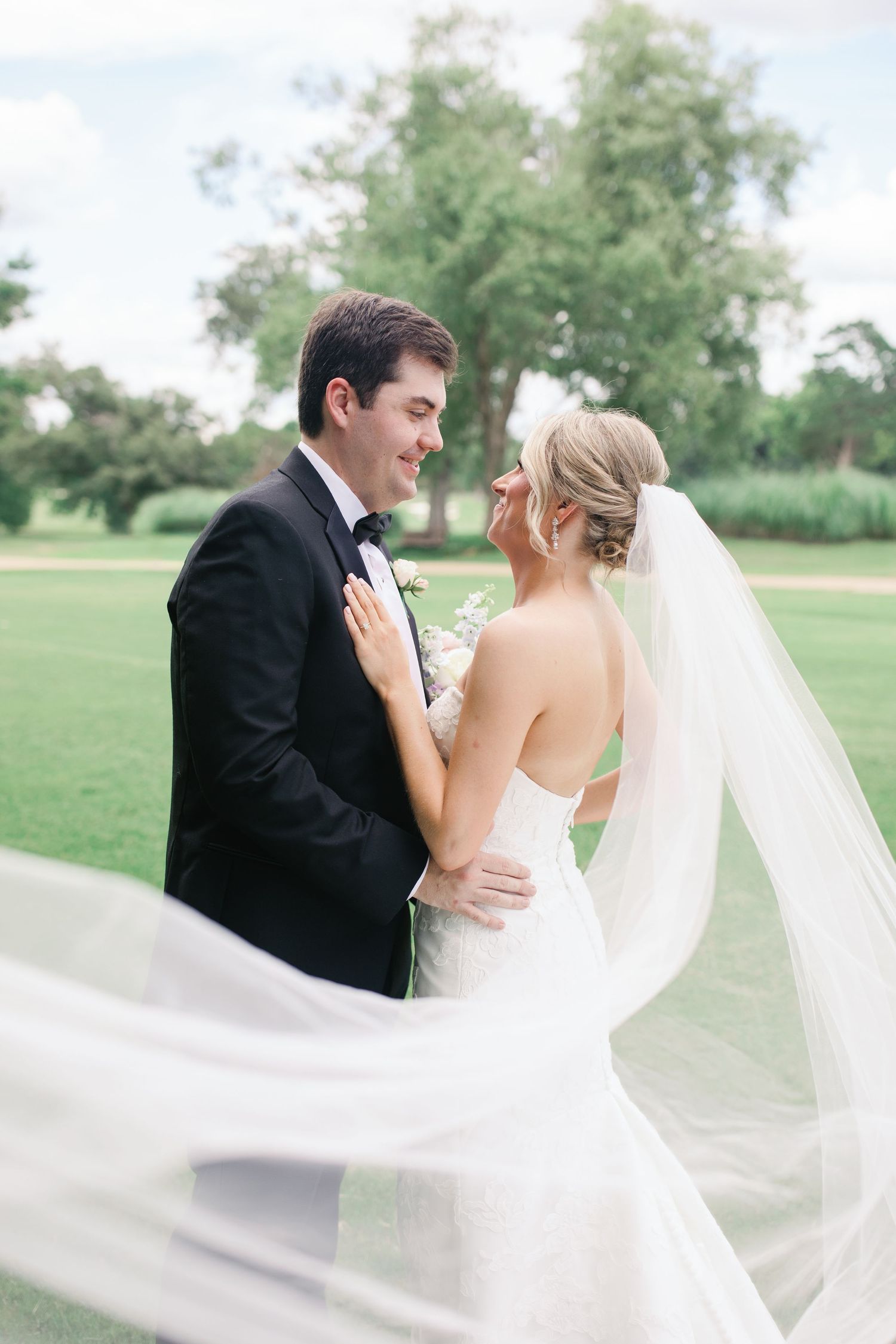 Bride and groom share a romantic moment outdoors, her veil flowing in the wind, surrounded by lush green trees.
