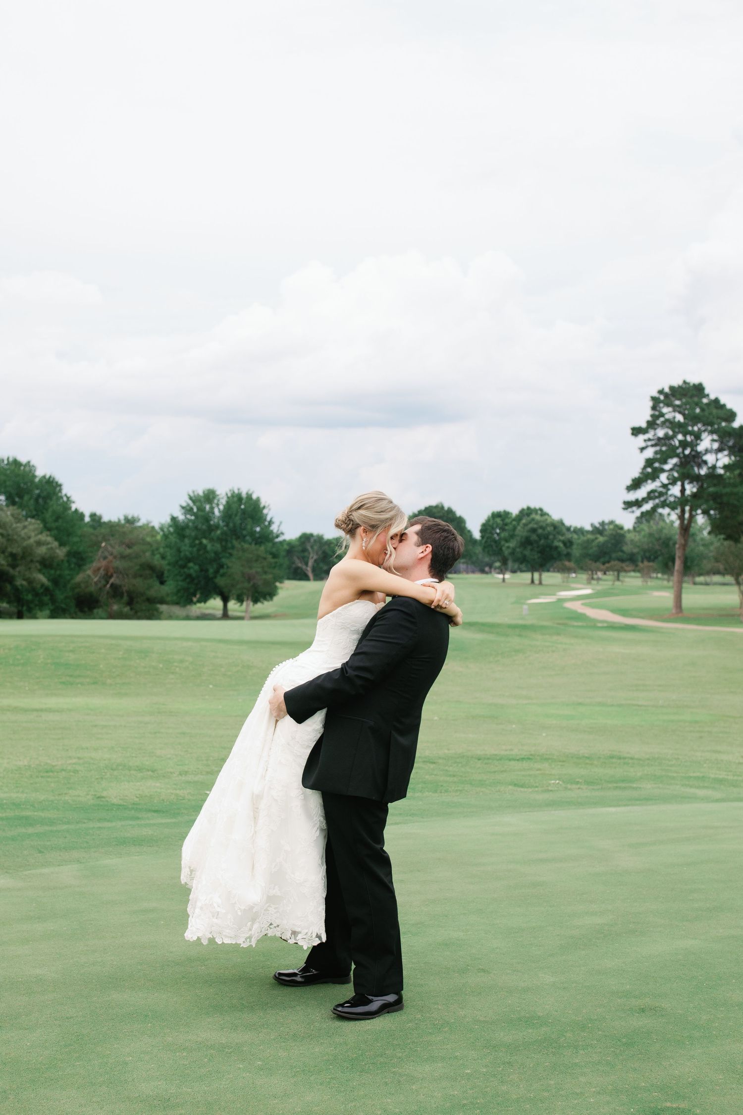 Groom lifts bride in white wedding gown for a romantic kiss on a lush green golf course under cloudy skies.