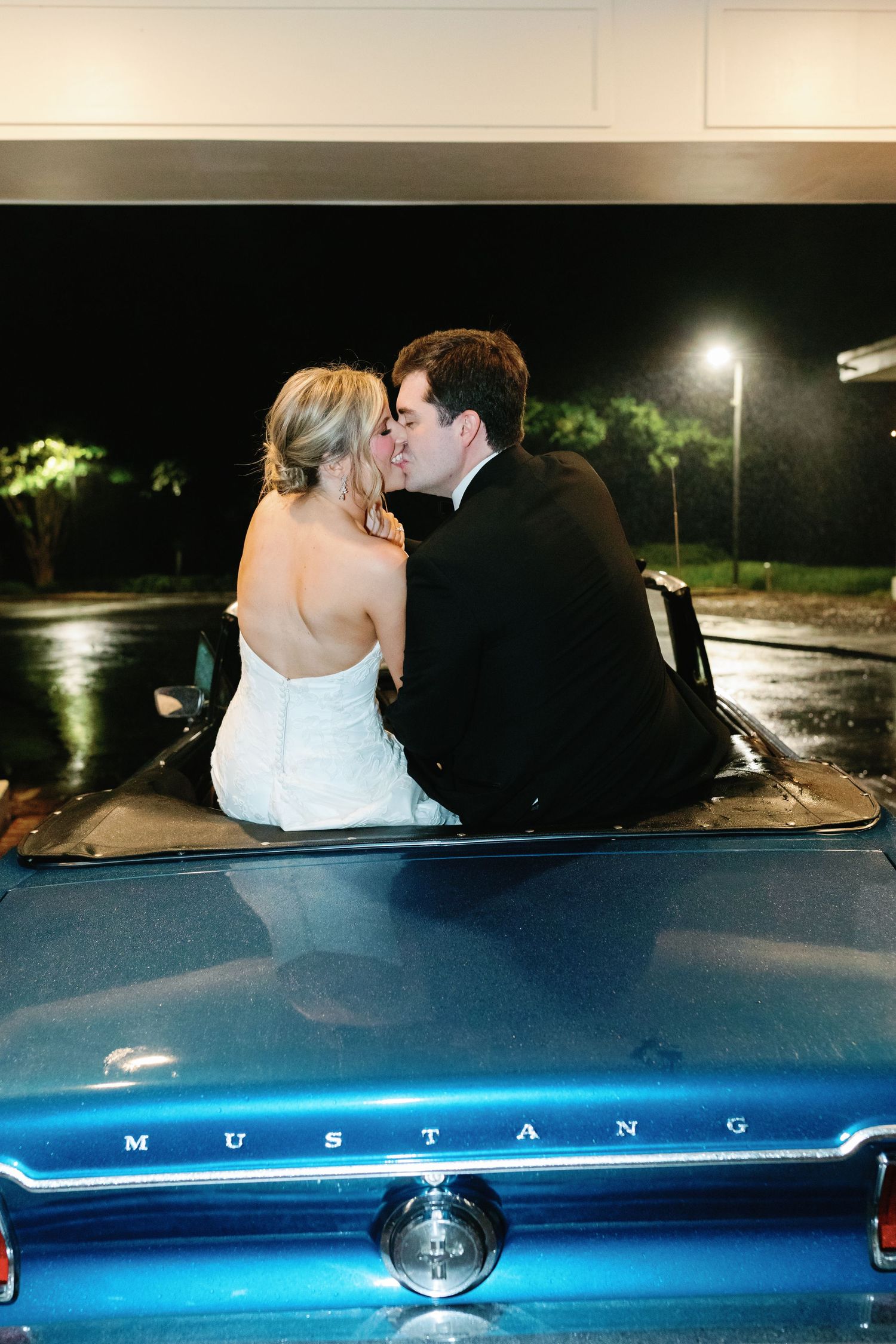 Bride and groom sharing a romantic kiss in a classic blue Ford Mustang convertible on their wedding night.
