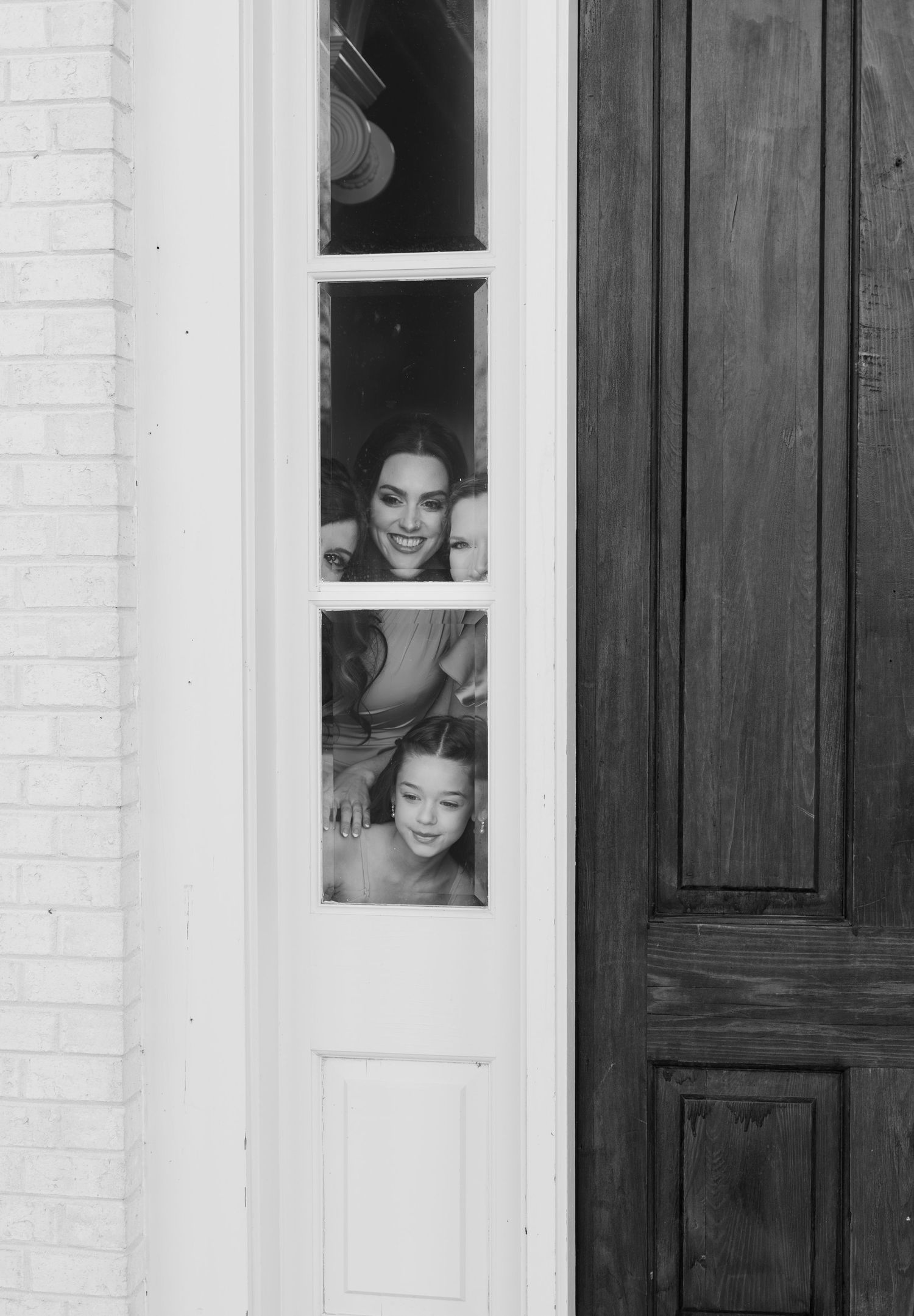 Two women smiling and peeking through a narrow sidelight window beside a dark wooden door in black and white.
