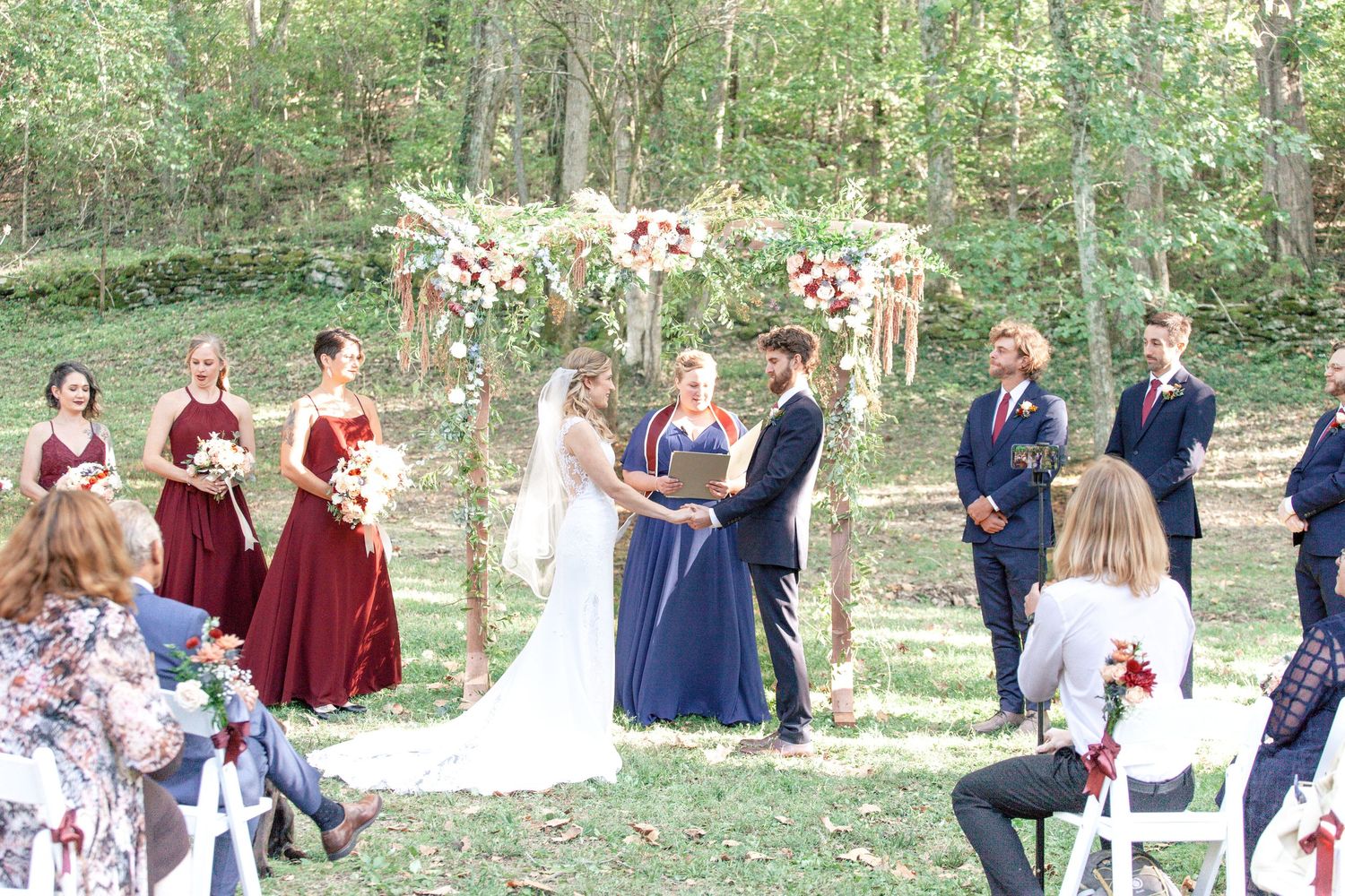 An outdoor forest wedding ceremony with burgundy-dressed bridesmaids and floral arch.