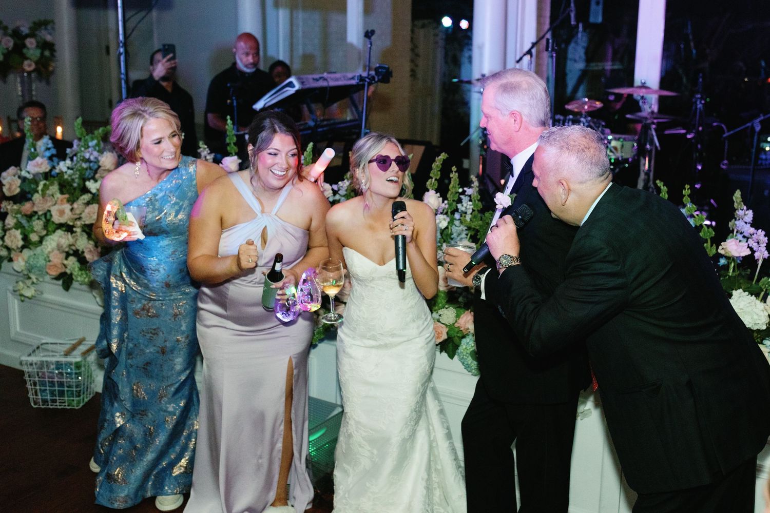 Bride in white gown singing with guests at wedding reception, surrounded by floral arrangements and band.