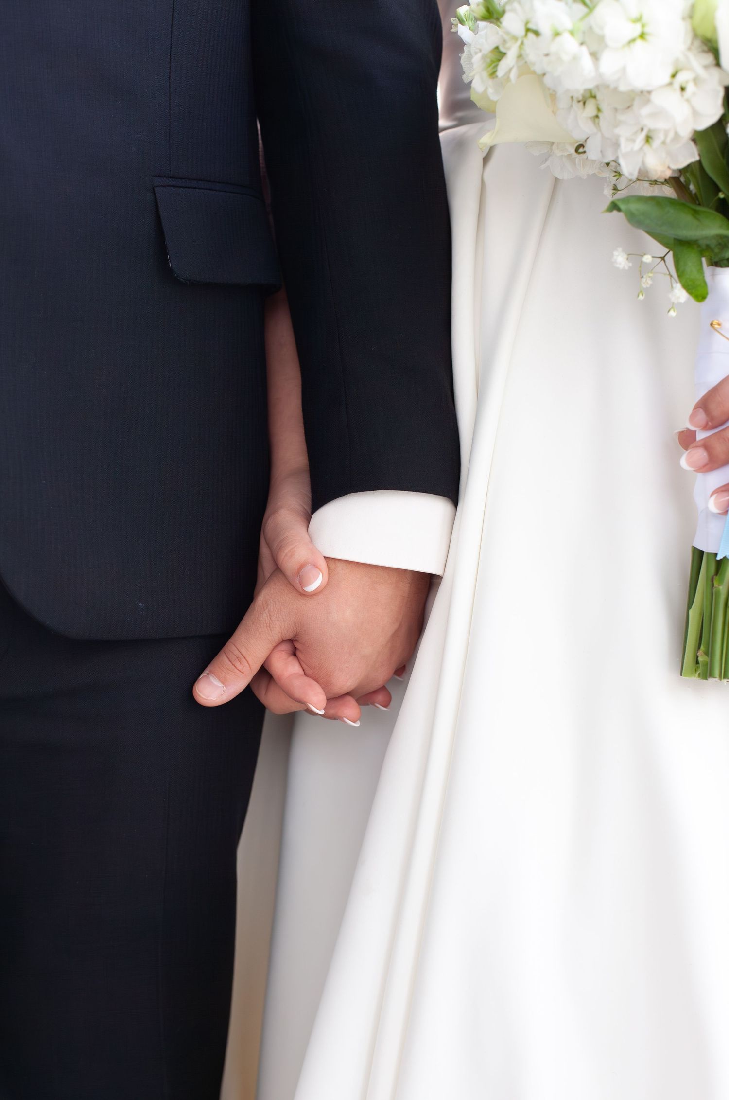 Bride and groom holding hands at wedding, white bridal gown with bouquet of white flowers and dark navy suit.