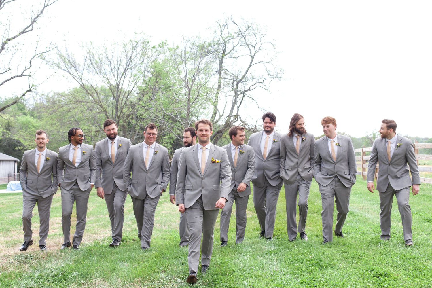 Groom leads groomsmen in matching gray suits across green lawn at outdoor wedding venue.