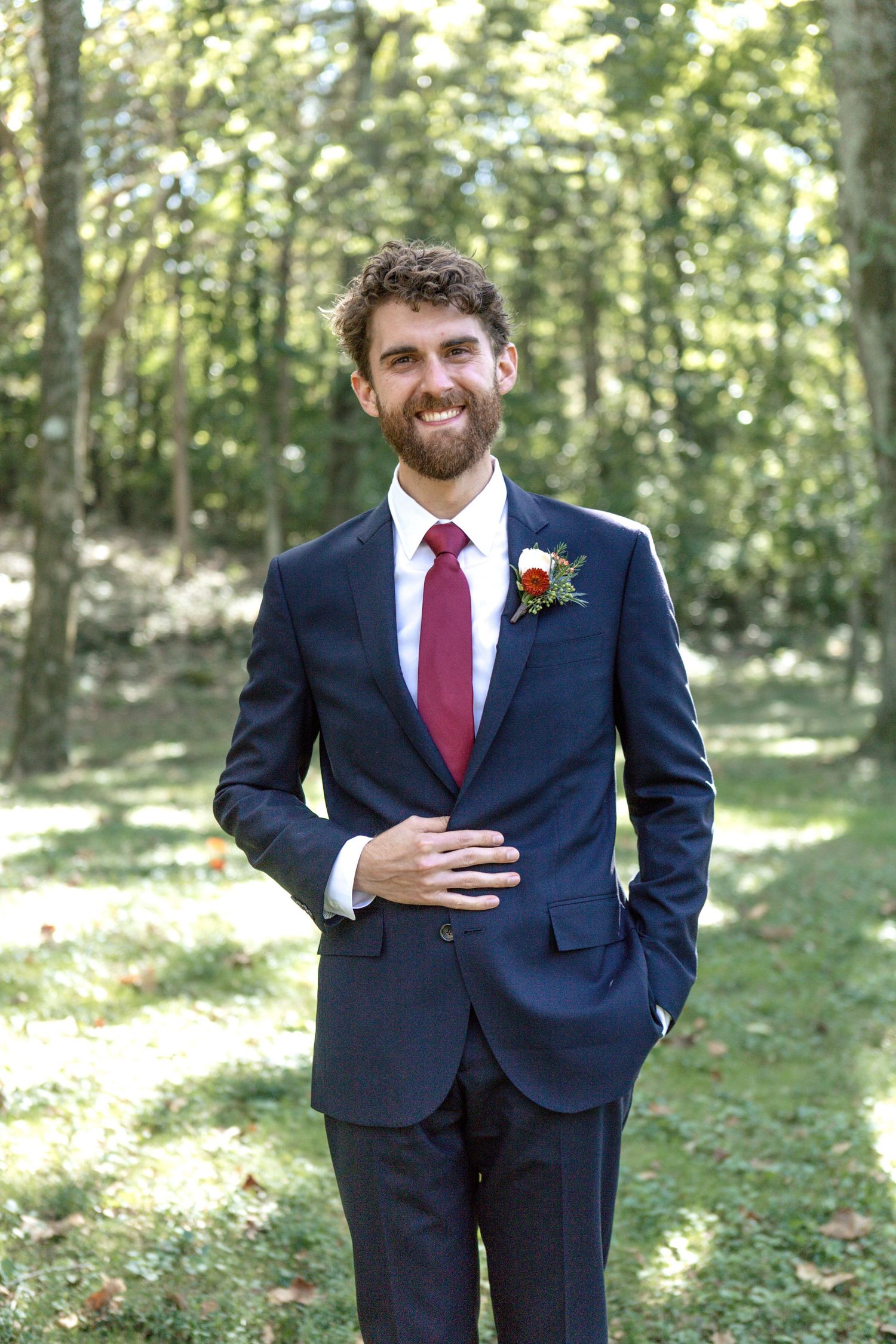 A groom in navy suit with burgundy tie poses in a sunlit forest setting.