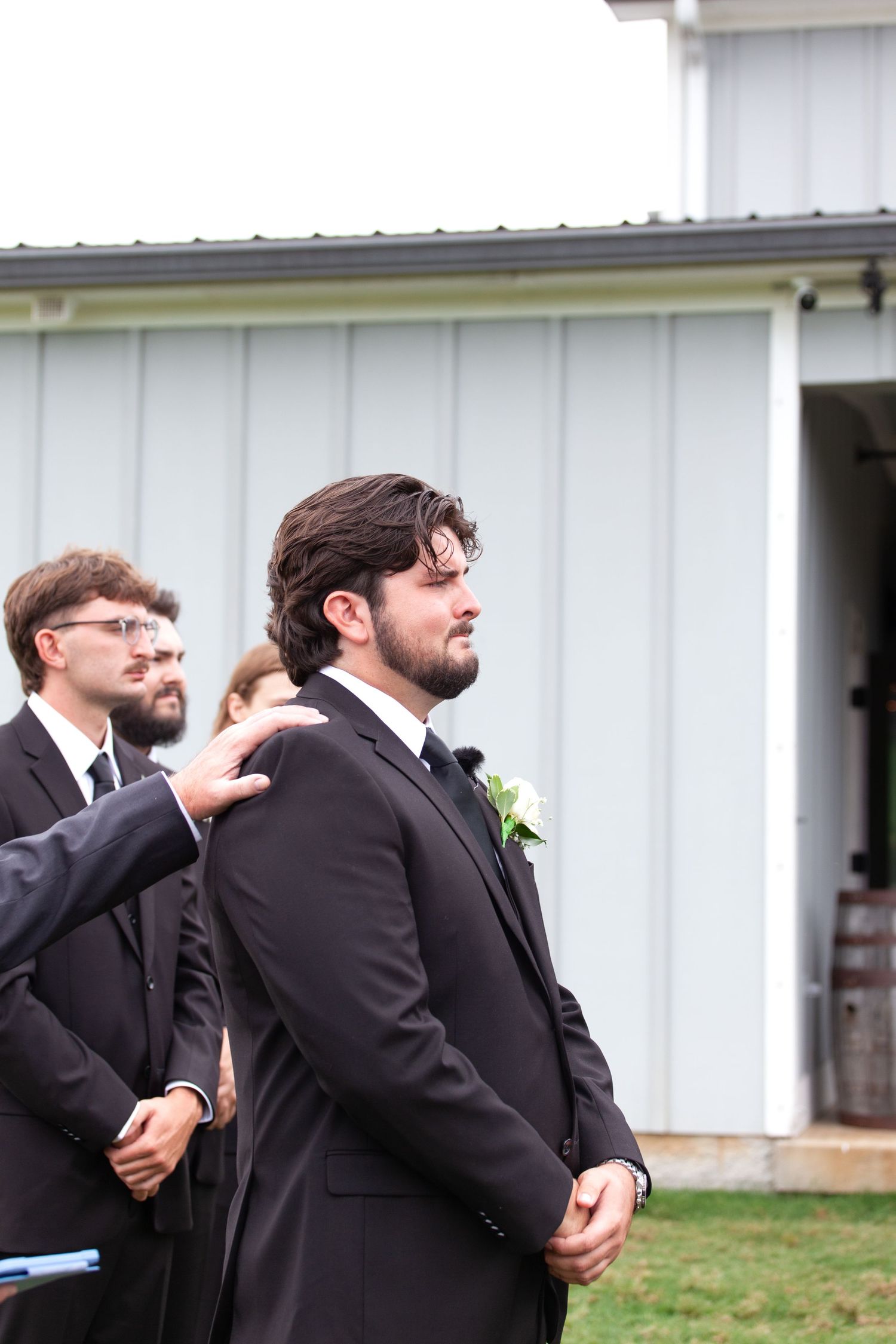Groom in black suit with boutonniere stands emotionally at outdoor wedding ceremony near white barn.