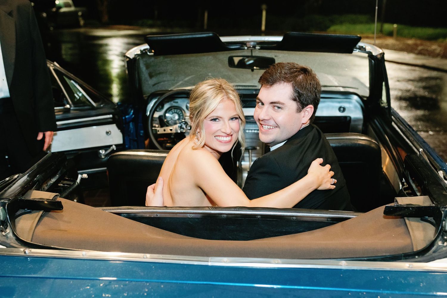 Couple smiling in a classic blue convertible car, woman in strapless dress embracing man in suit.