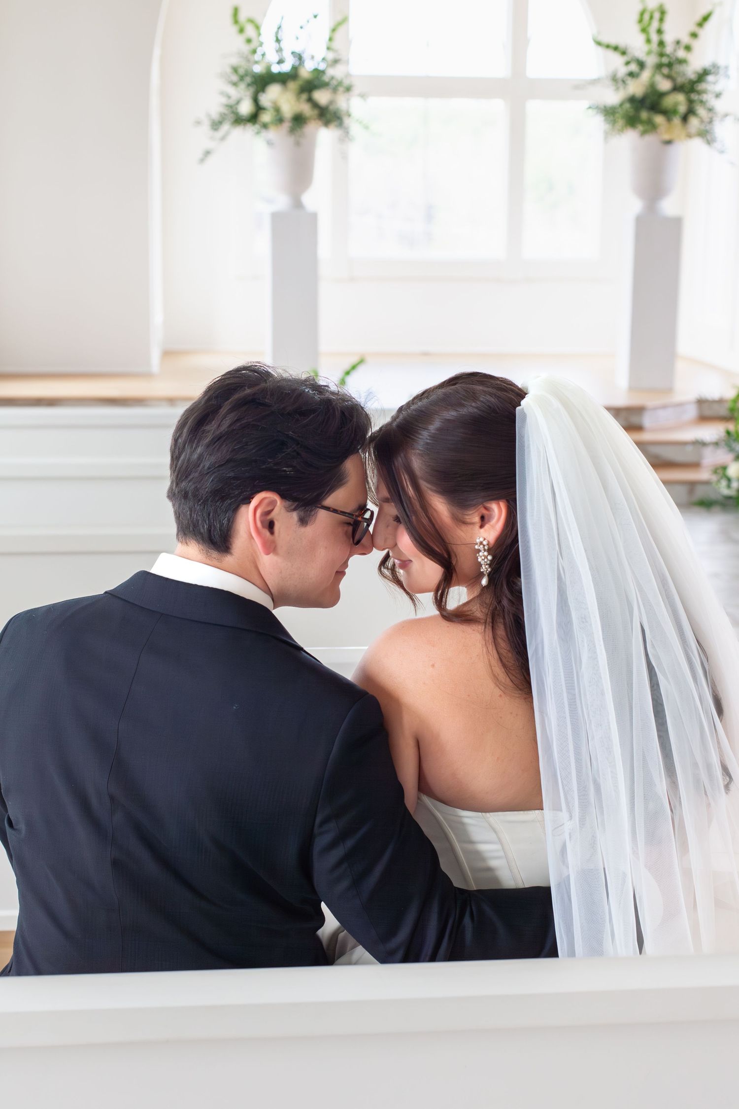 Bride and groom share an intimate moment touching foreheads while seated in a sunlit white church.