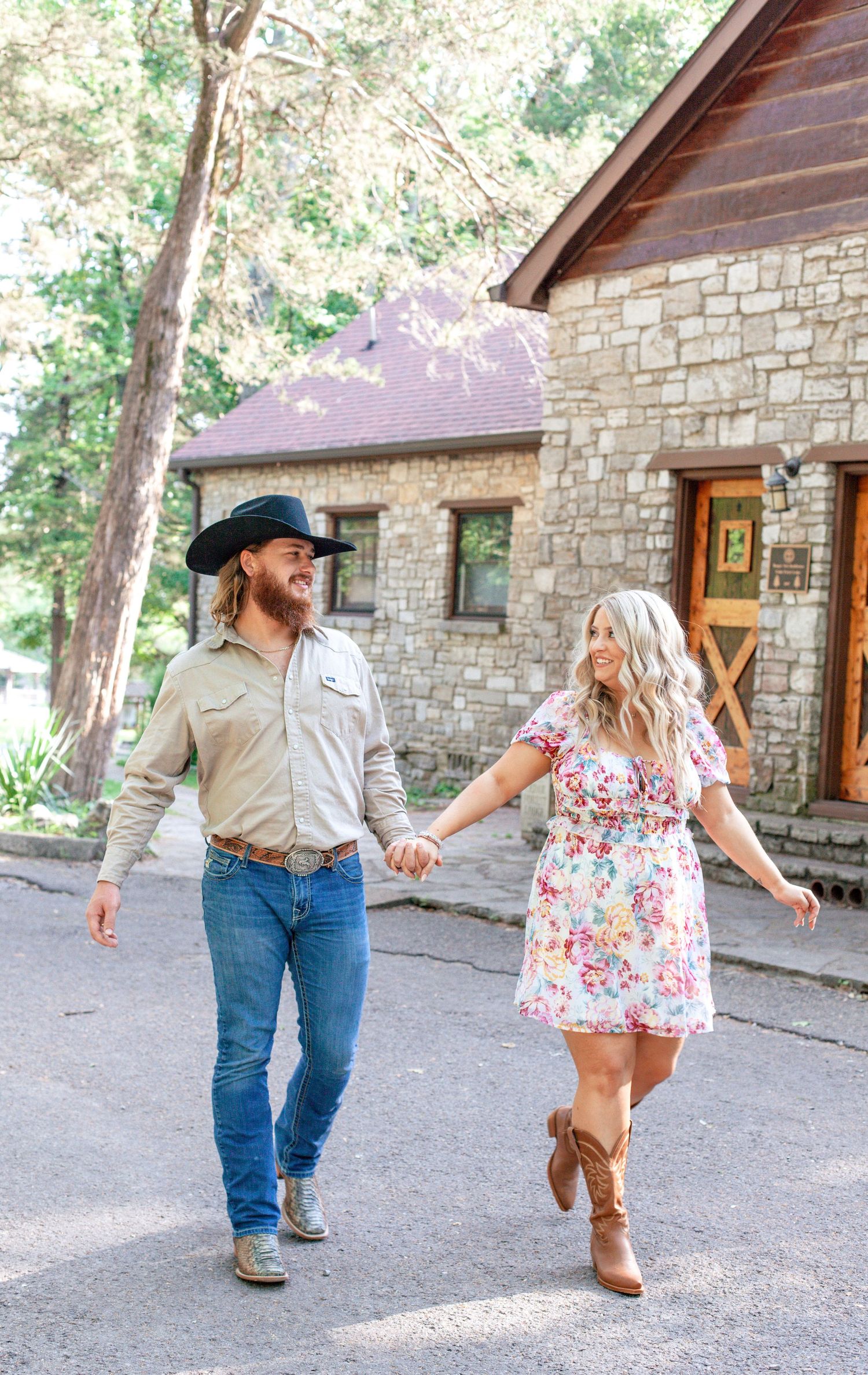 A couple in casual attire dances together on a gravel path outside a rustic stone building on a sunny day.