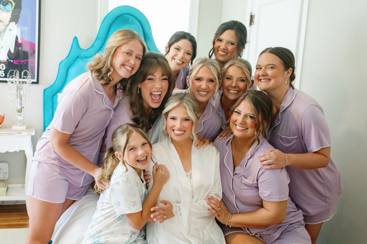 Bride in white robe smiles with bridesmaids in purple pajamas and flower girl during wedding morning preparations.