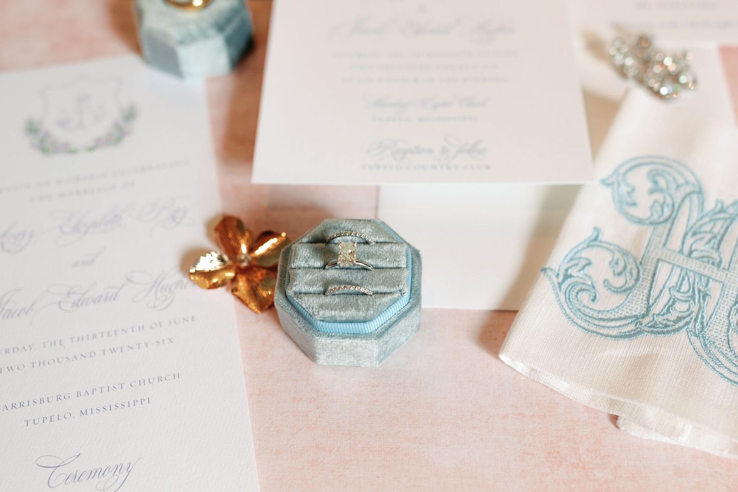 Elegant wedding flat lay featuring a blue velvet ring box, monogram stationery, and gold bow on a soft pink background.