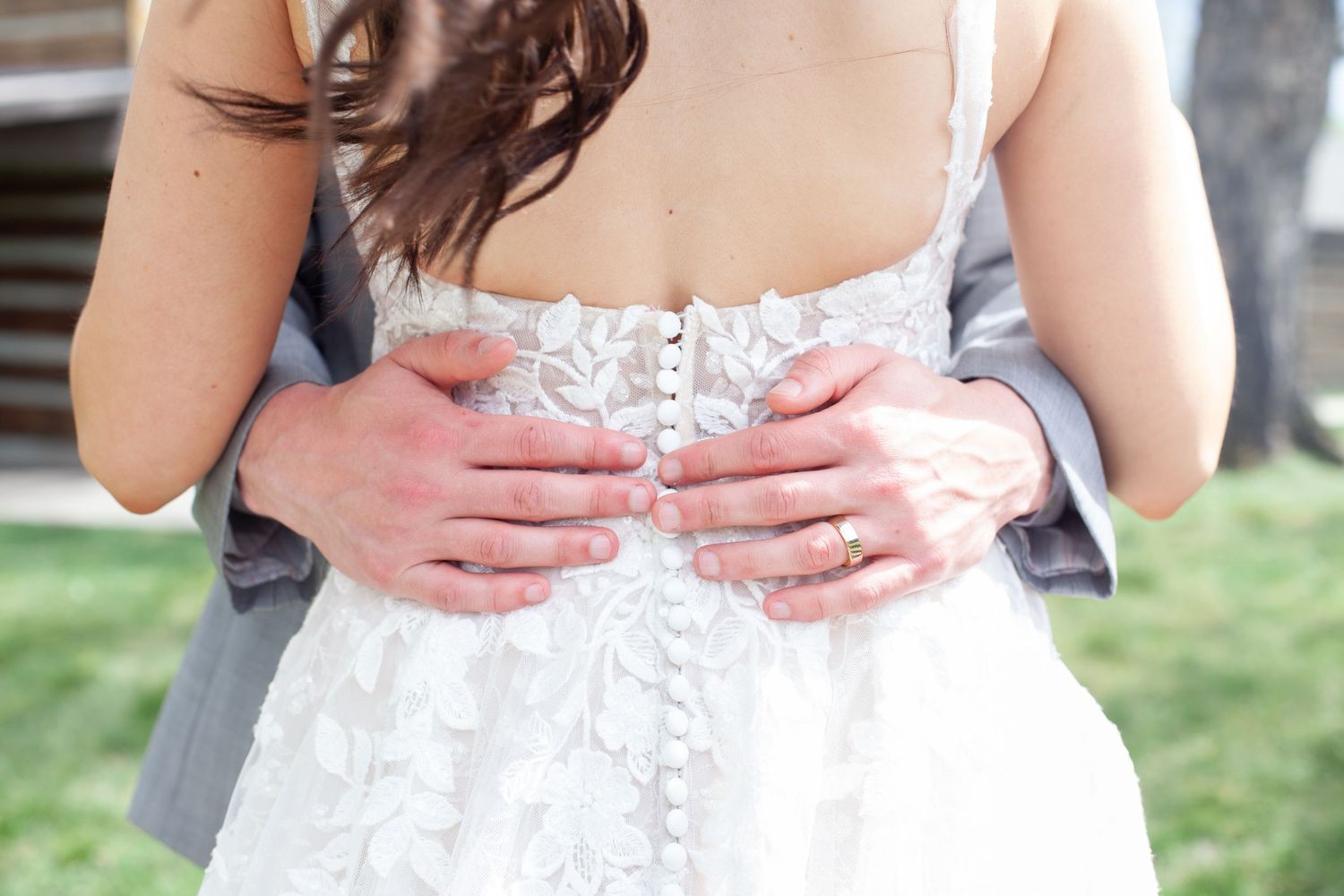 Groom in gray suit embracing bride in lace wedding dress from behind, showing button-back detail.