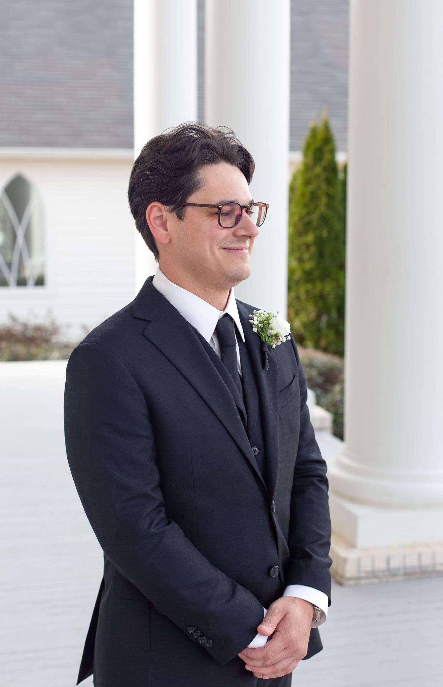 Smiling groom in dark suit with boutonniere and glasses standing outside elegant white venue.
