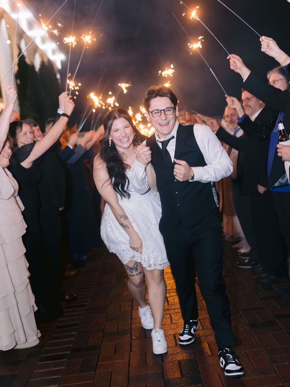 Newlywed couple exits wedding reception through sparkler tunnel, bride in white dress, groom in vest and glasses.