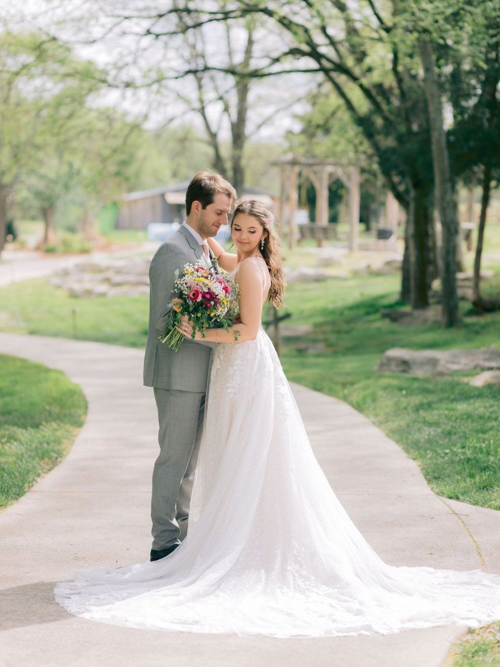 Bride and groom pose on garden path, she holds colorful bouquet in white gown, he wears gray suit, lush greenery behind.