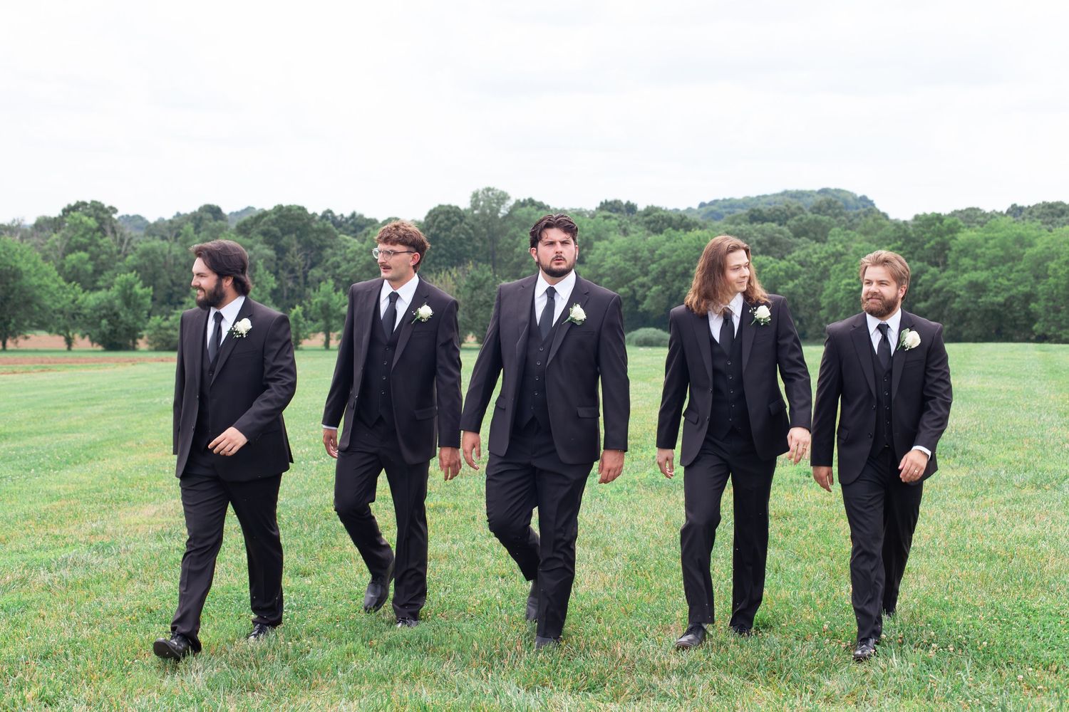 Five groomsmen in black suits with white boutonnieres walking across a green field at an outdoor wedding.