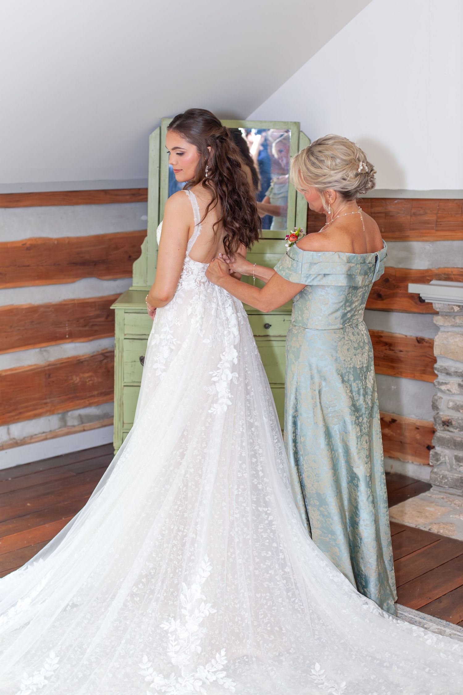 A bride in a lace wedding gown being helped by an older woman in a teal dress near a vintage mirror in a rustic log cabin.