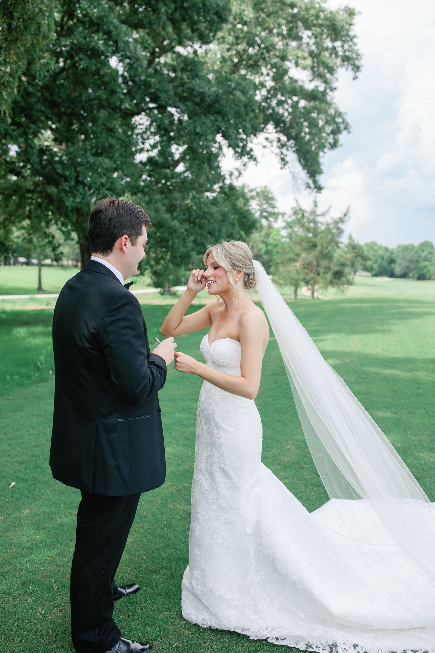 Bride wiping tears while groom holds her hands during emotional first look on lush green golf course.