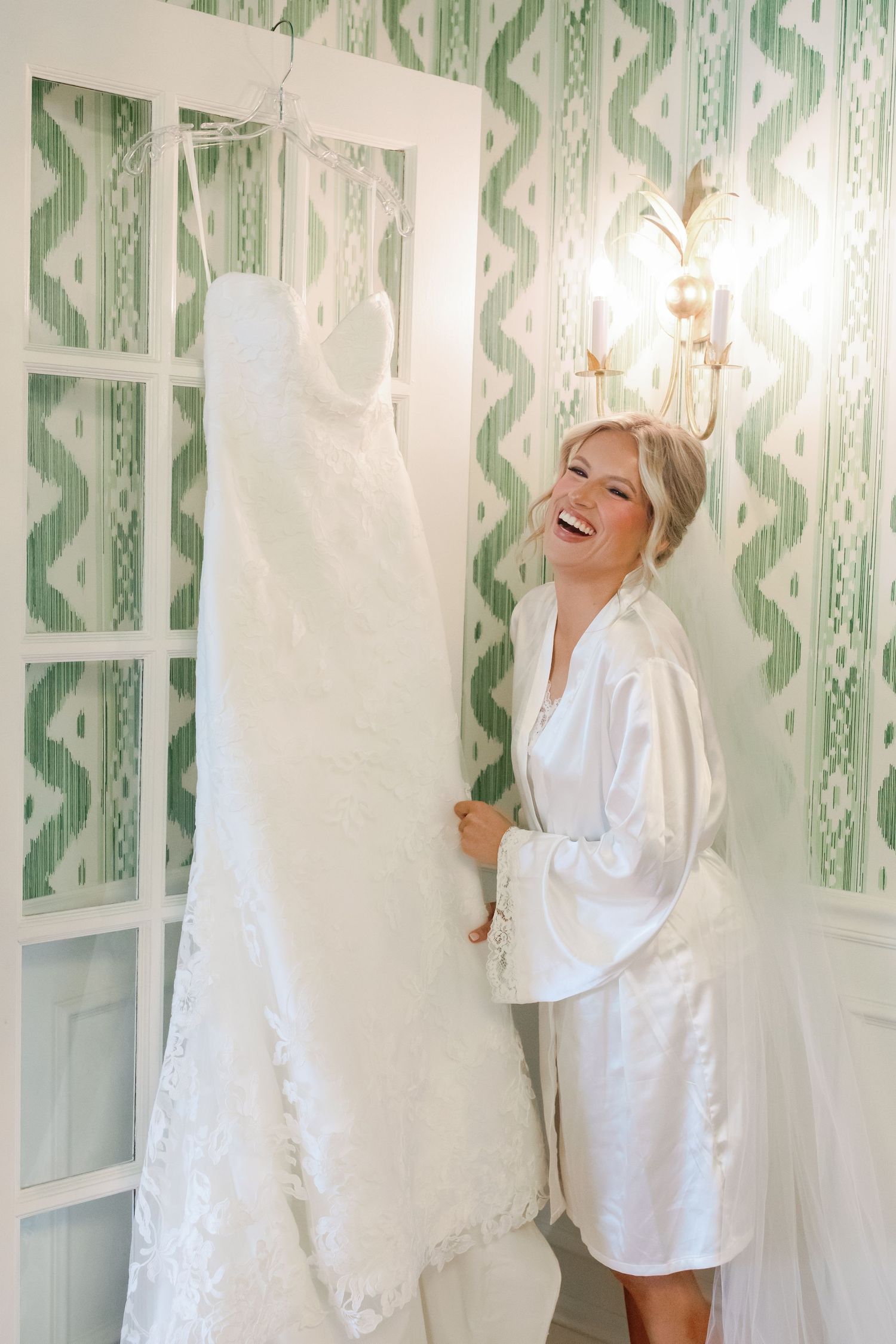 Smiling bride in white satin robe admiring her lace wedding dress hanging in a room with green patterned wallpaper.