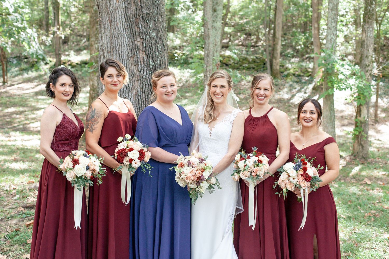Bride in white gown with five bridesmaids in burgundy and navy dresses holding floral bouquets outdoors among trees.