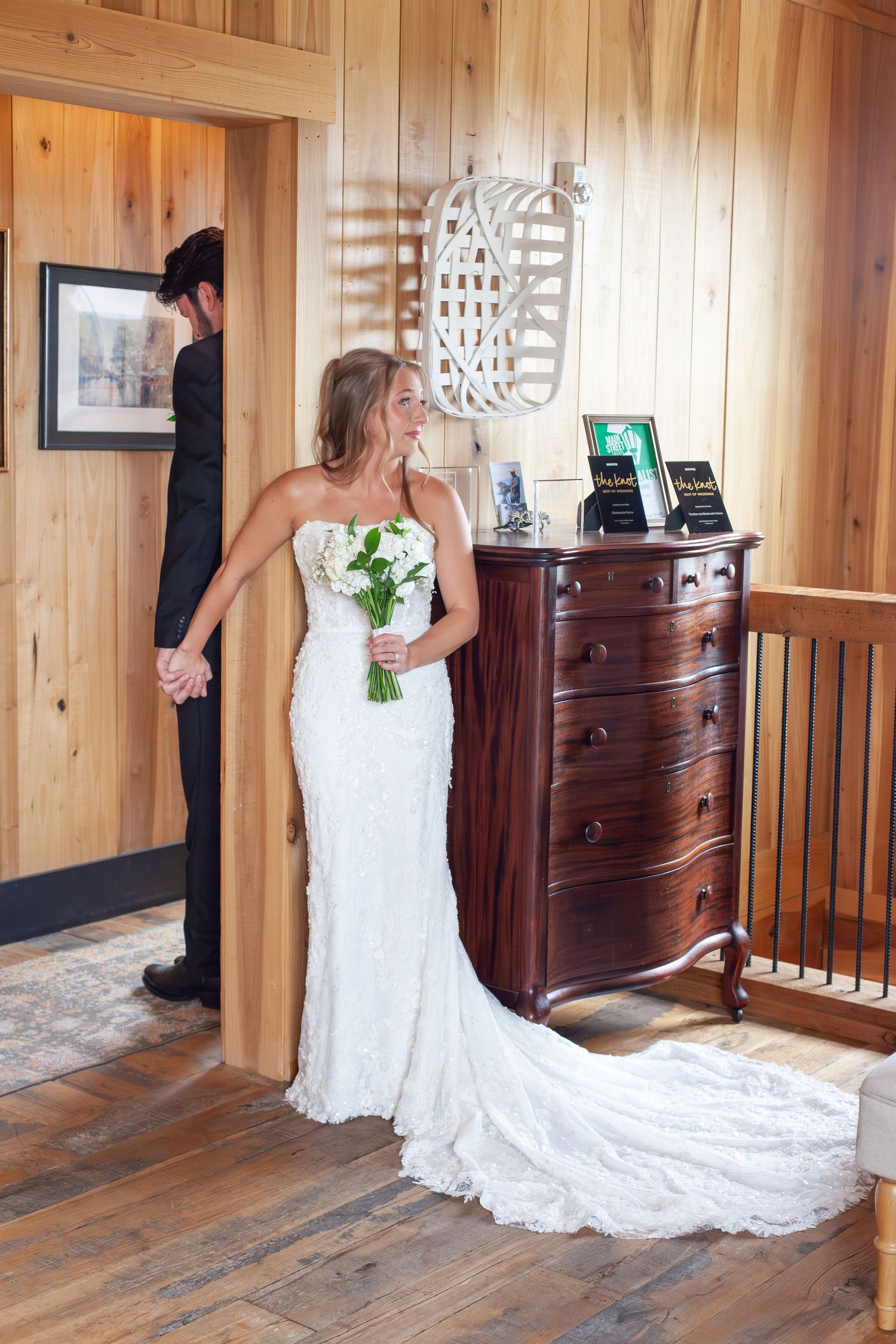 Bride in white lace gown holds bouquet during first look moment with groom in rustic wood-paneled venue.