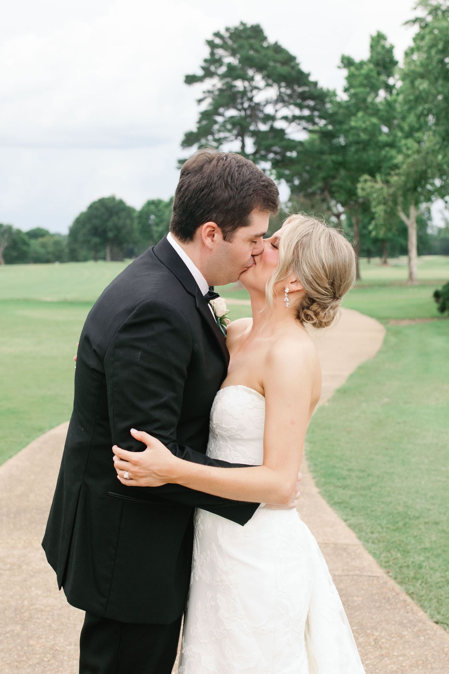 A bride and groom share a romantic kiss outdoors on a golf course pathway, dressed in a white gown and black tuxedo.