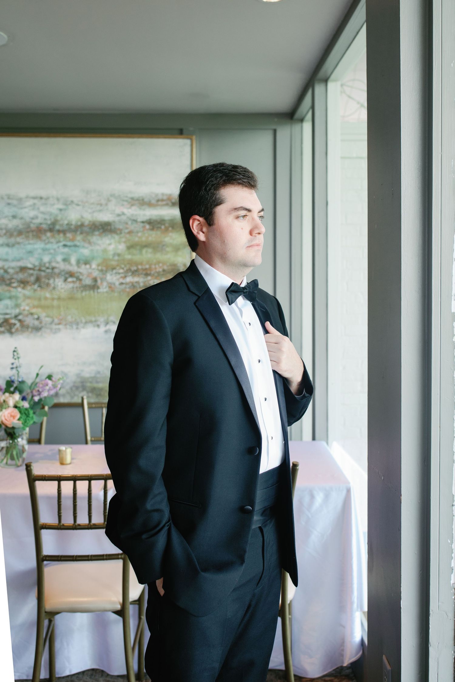 Groom in black tuxedo adjusting bow tie by window in elegant wedding venue.