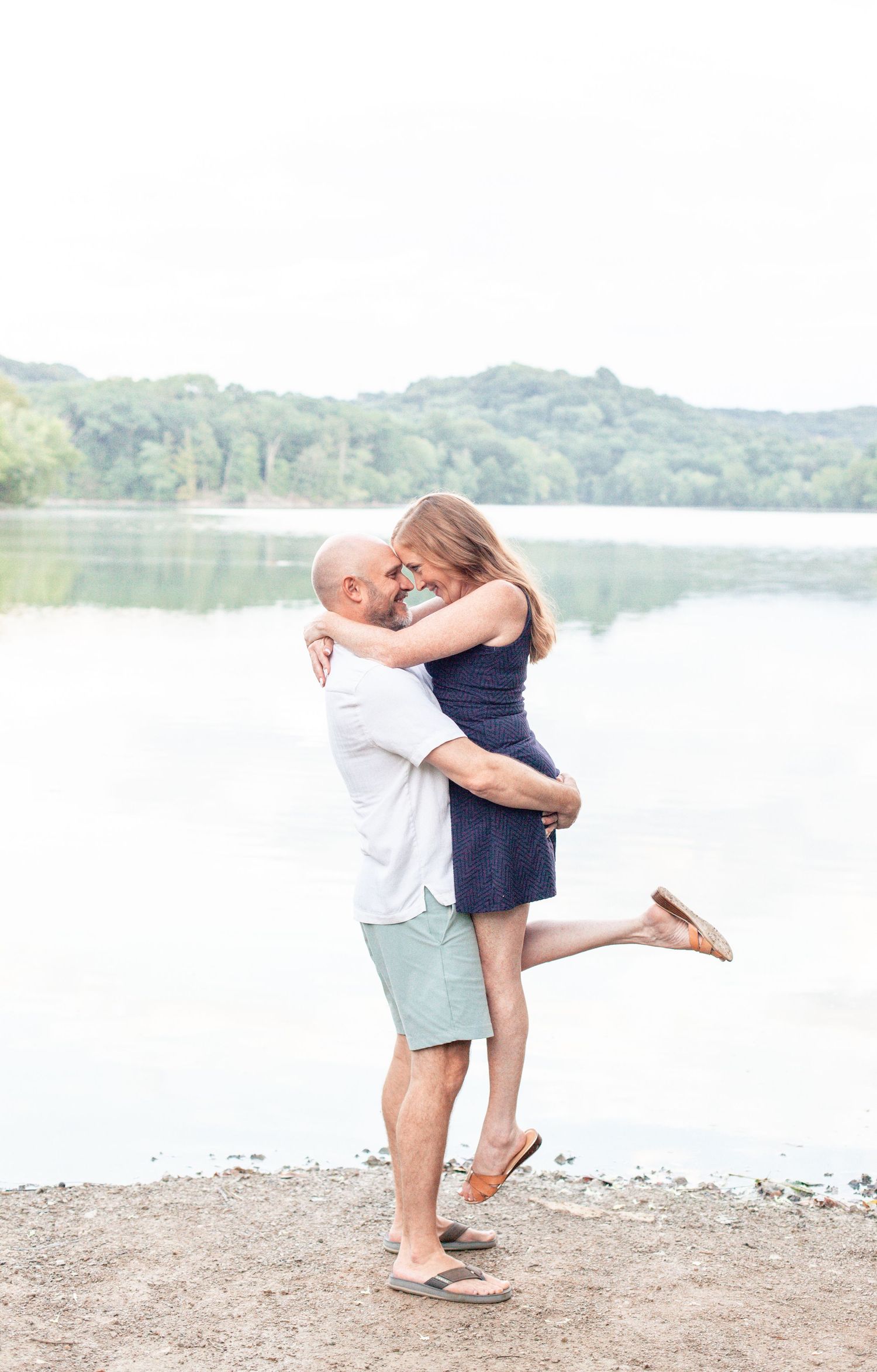 A couple embraces in a romantic moment by a serene lake, with green hills in the background.