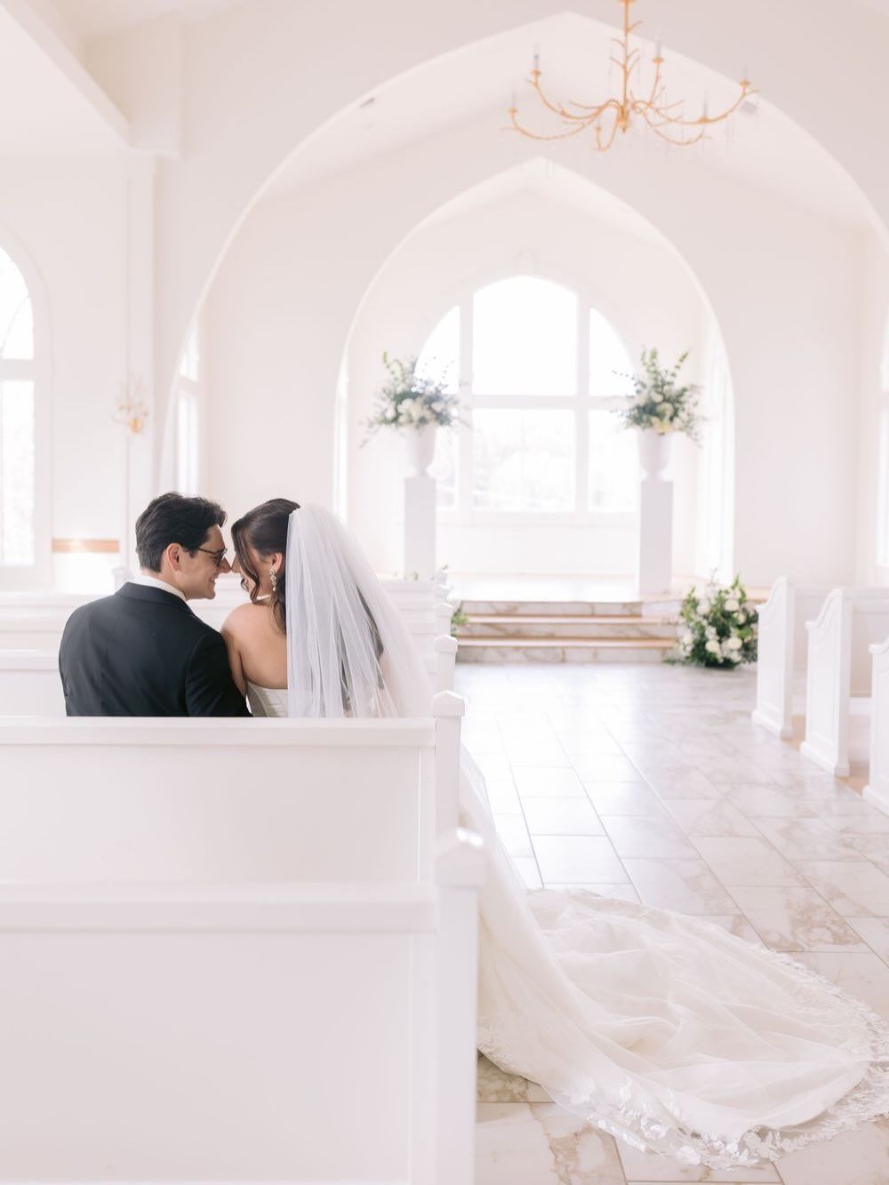 Bride and groom share an intimate moment in a stunning all-white chapel with arched ceilings and floral altar arrangements.
