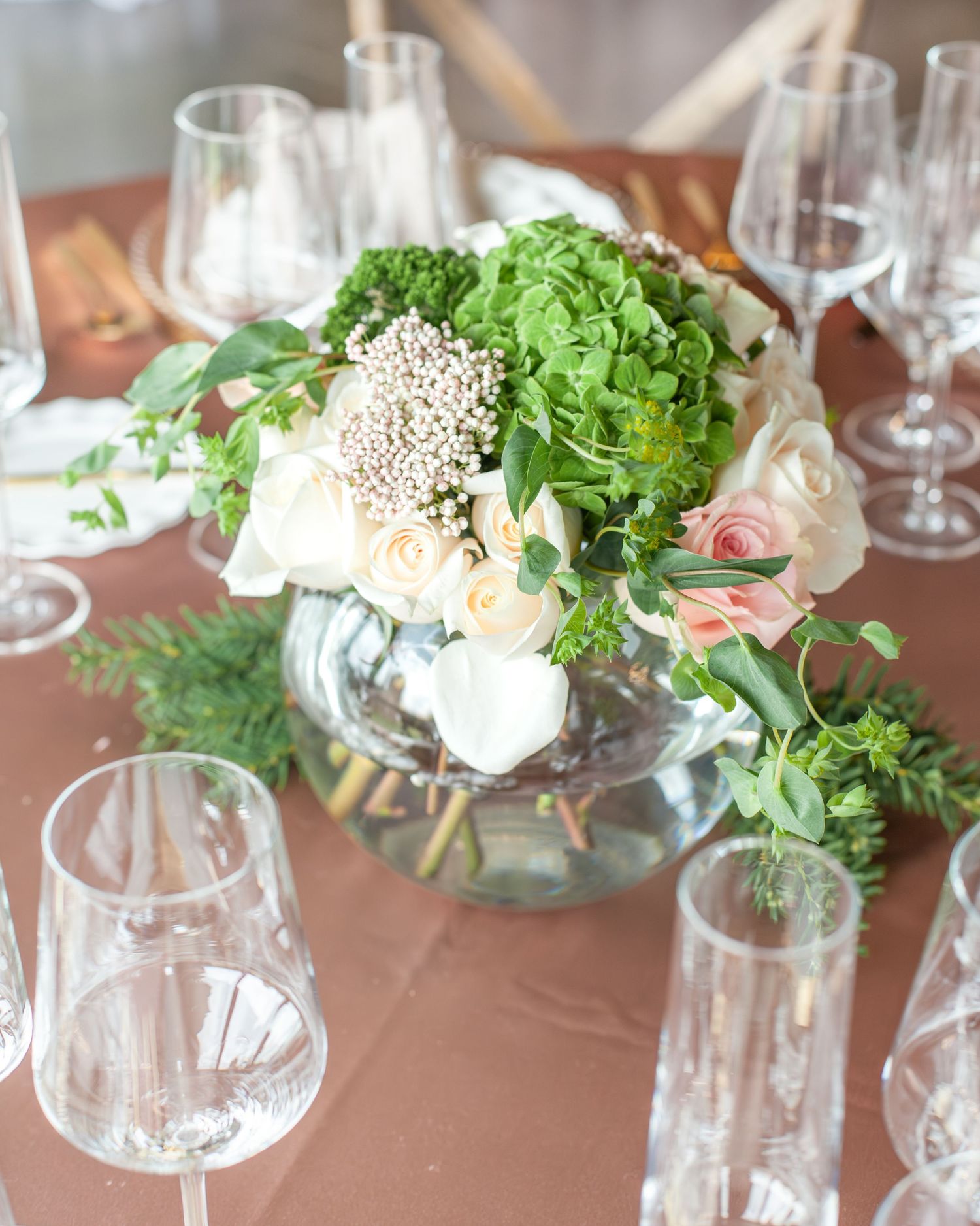 Elegant wedding centerpiece with white roses, green hydrangeas, and eucalyptus in a glass bowl surrounded by crystal glasses.